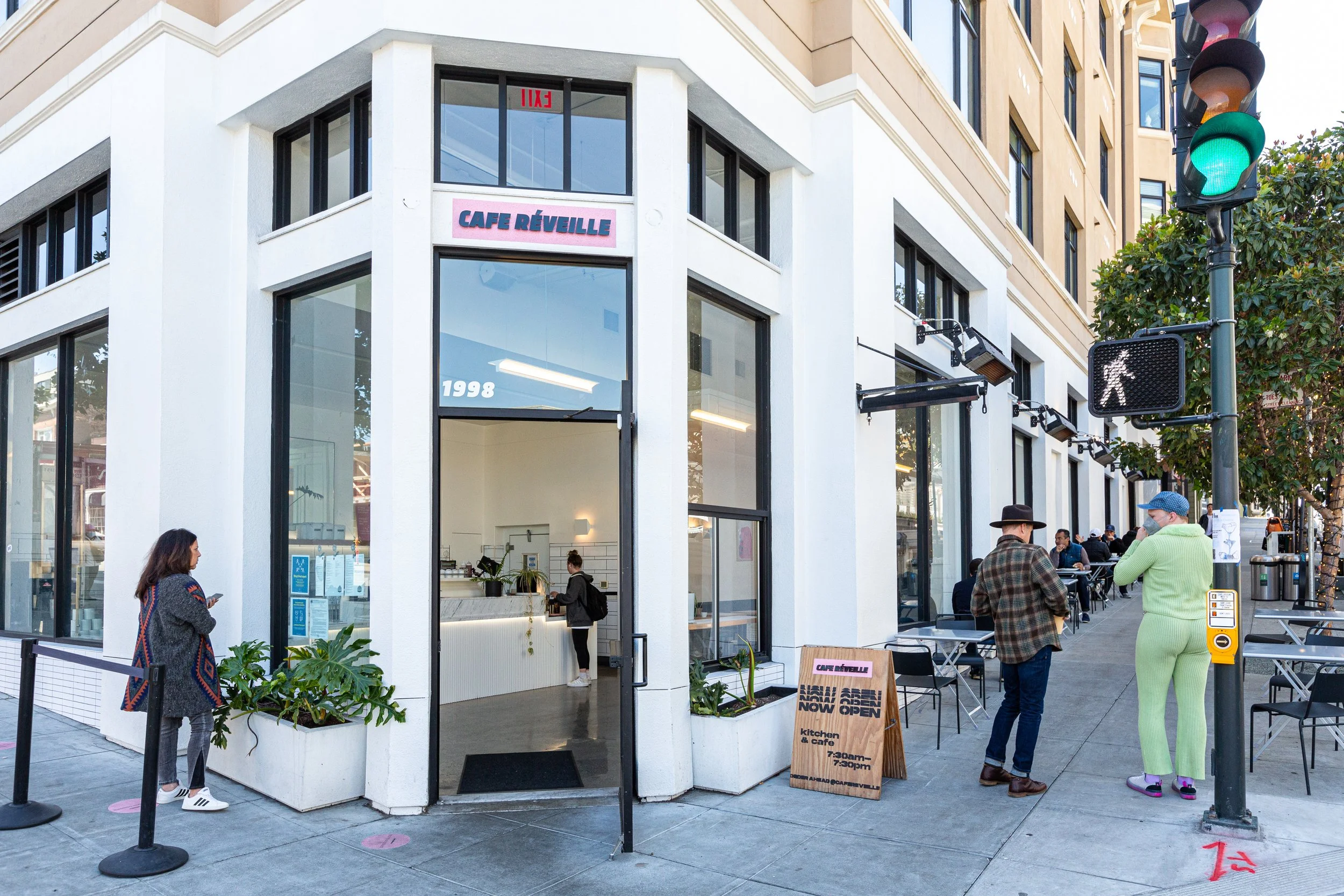 A corner storefront of Cafe Réveille on a city street with people waiting outside. The cafe has large glass windows and a pink sign with names in blue. There are outdoor seating areas and pedestrians, including a woman in a patterned coat and white s