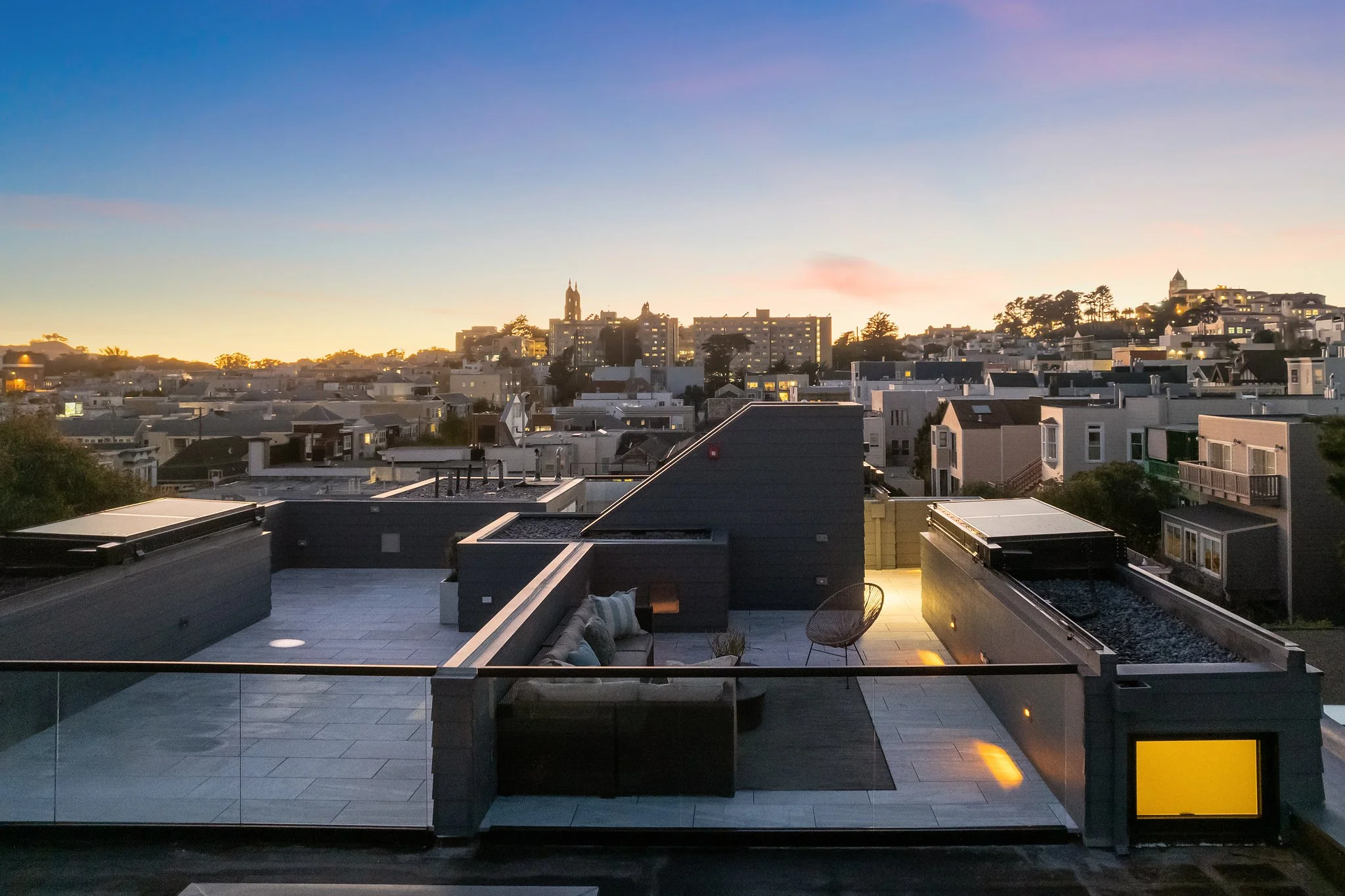 A city skyline view at sunset from a rooftop terrace showing residential buildings, some with lit windows, and a distant church steeple with a sky that transitions from yellow near the horizon to blue and purple higher up.