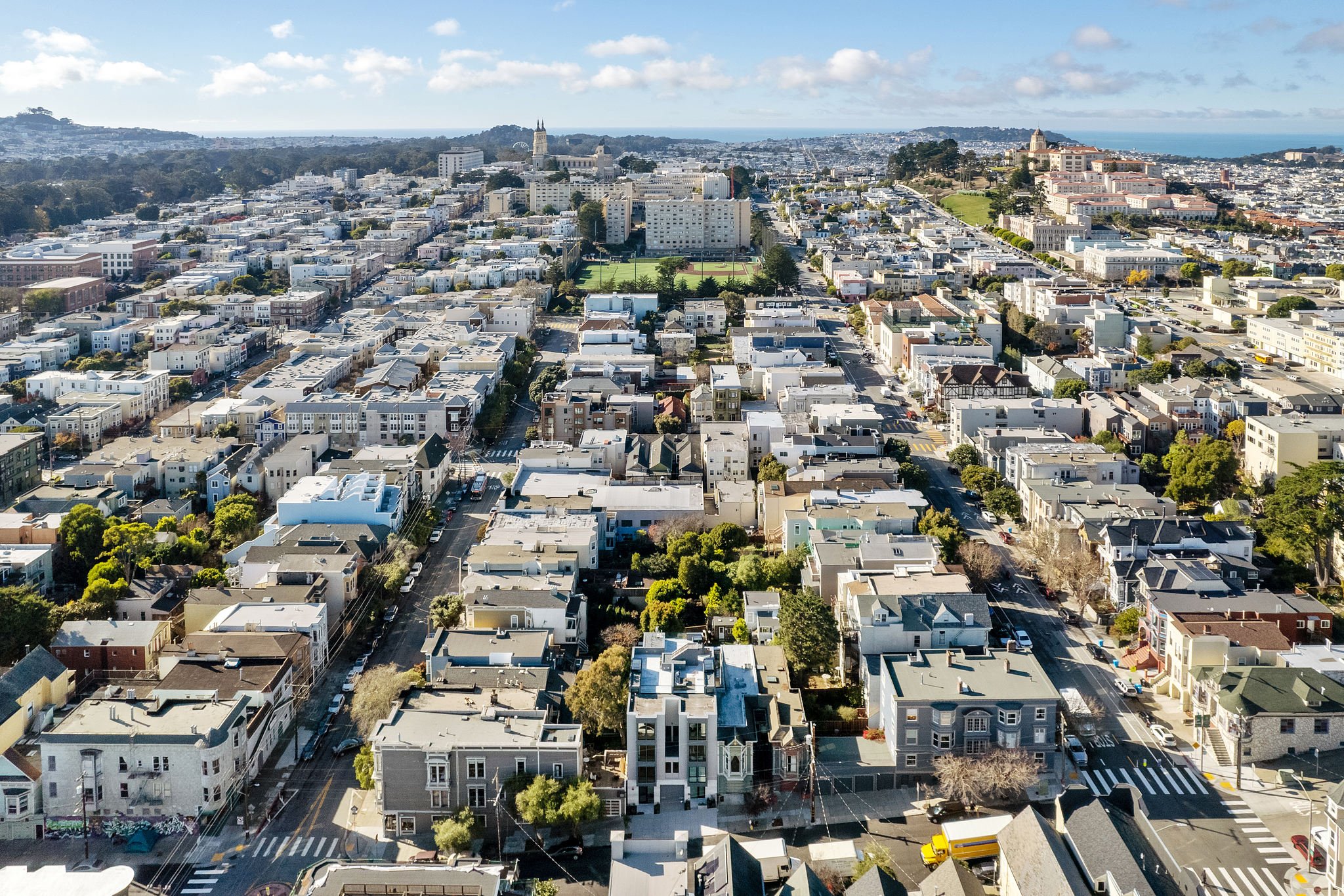 Aerial view of a city with tightly packed white and beige residential buildings, a green sports field, and distant hills with a church tower and the coastline visible in the background.