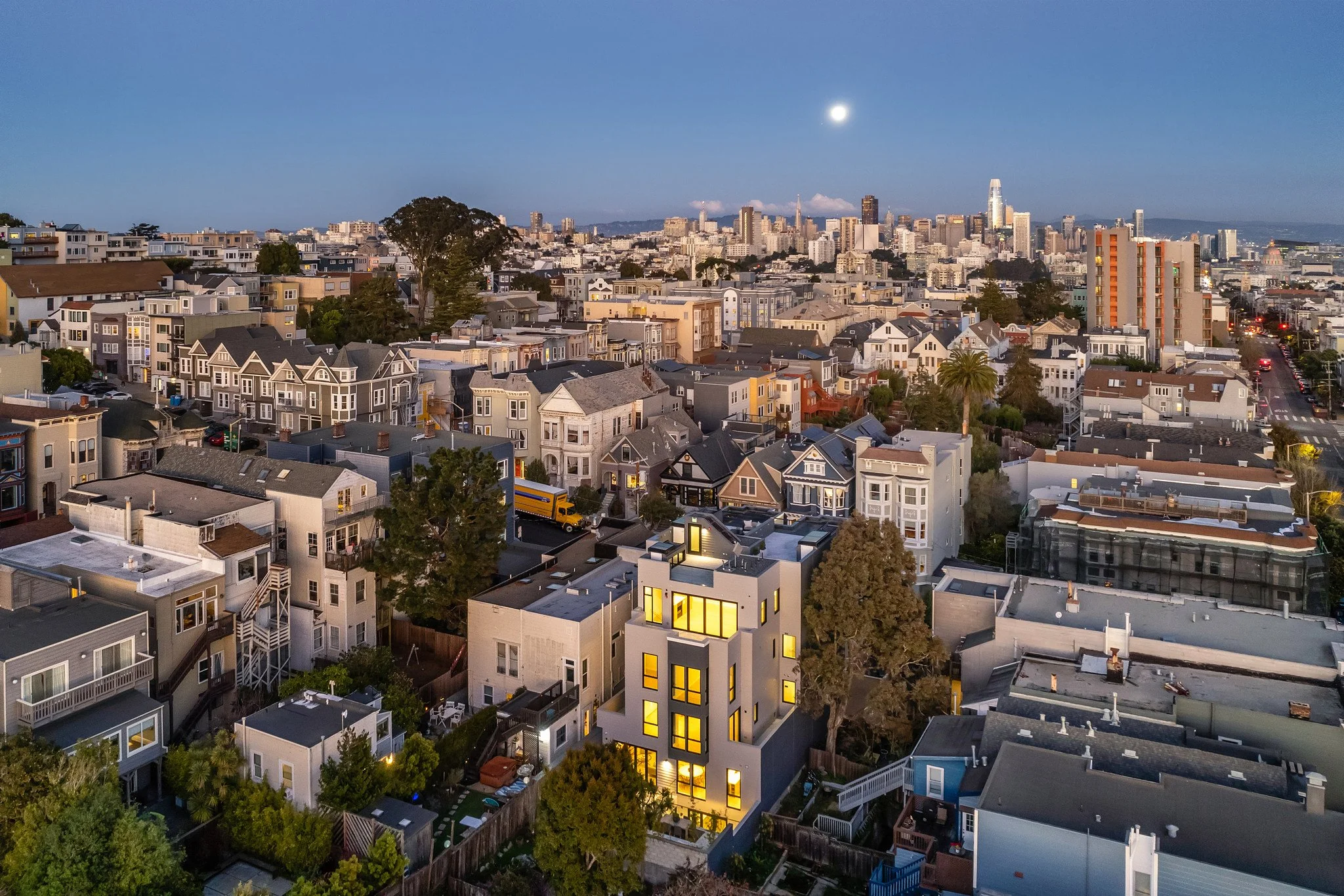 A panoramic view of a cityscape at dusk with numerous residential houses in the foreground, a cluster of high-rise buildings in the background, and a clear sky with the moon visible.