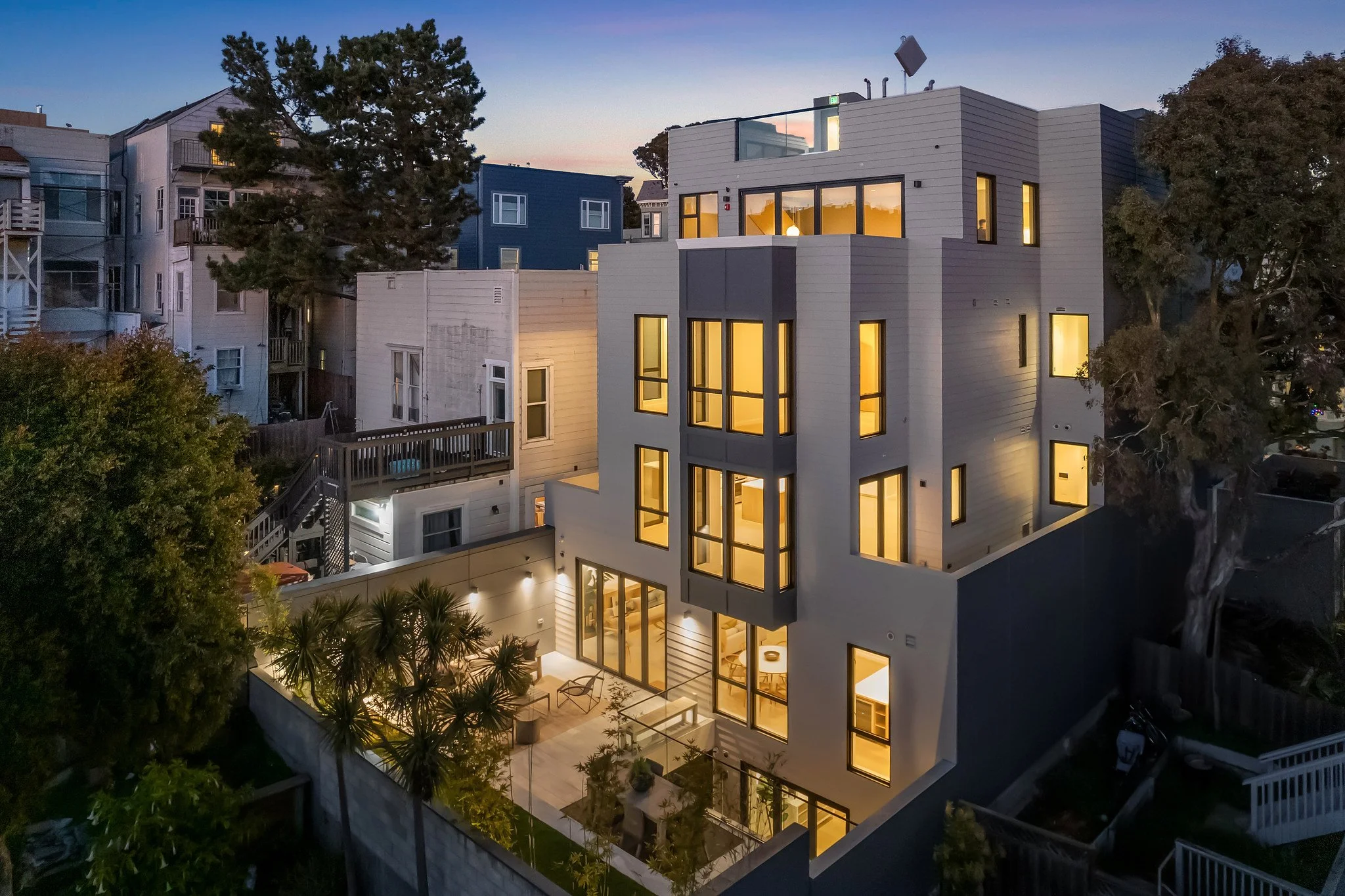 A modern multi-story house with large windows glowing with warm light, surrounded by trees and neighboring houses during sunset.