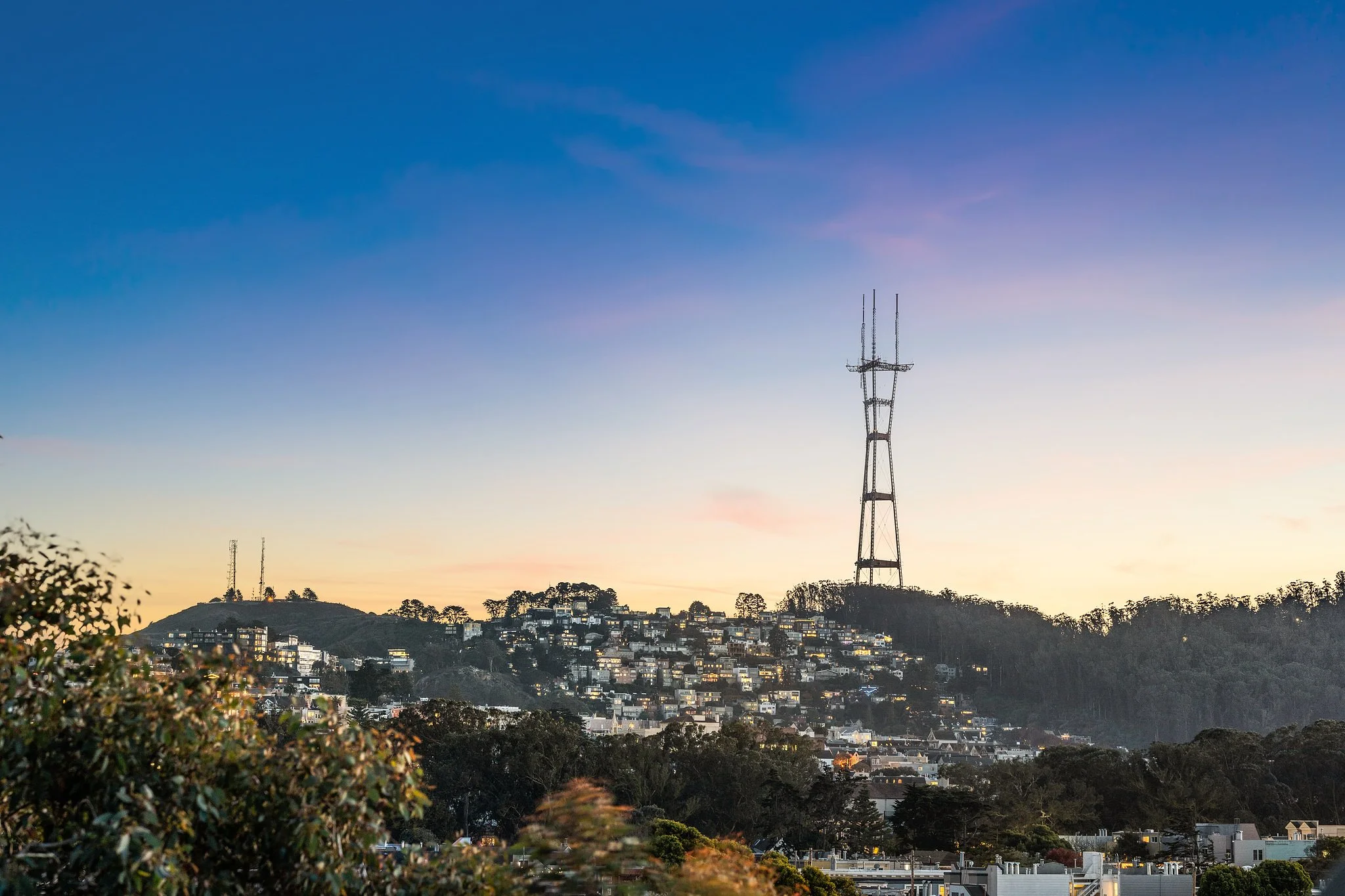 Cityscape during sunset with hills, residential buildings, and communication tower.