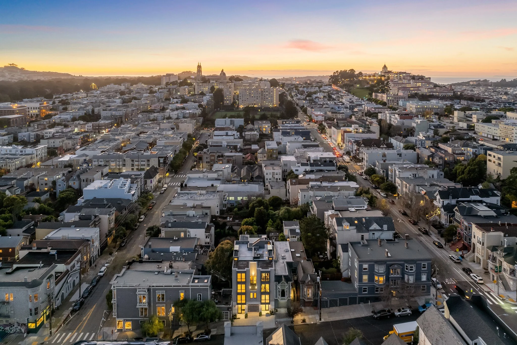 A cityscape at sunset with numerous houses, streets, and a few taller buildings. The sky has a warm gradient of colors from yellow to purple, and some buildings have their lights on.
