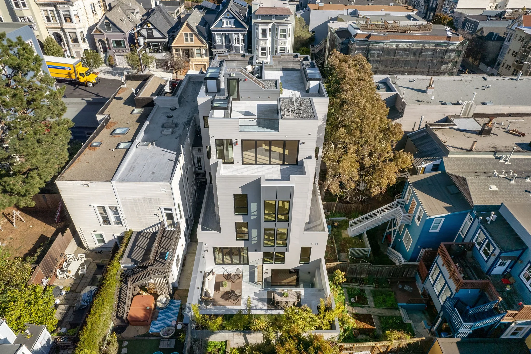 Aerial view of a modern multi-story house with a flat roof, large windows, and a backyard patio with furniture and trees, surrounded by neighboring houses in an urban residential area.