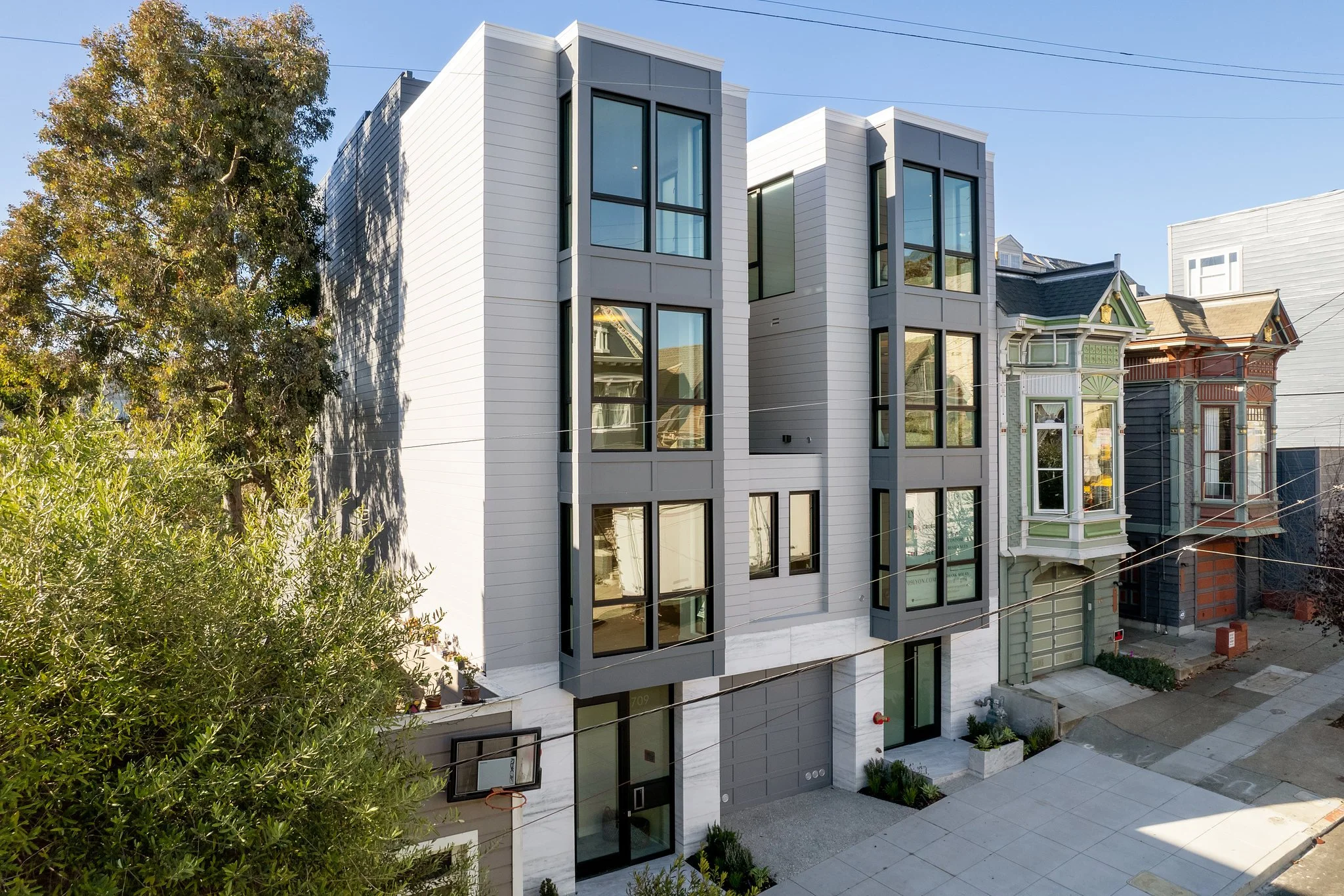 Modern multi-story building with large windows and gray exterior, located in an urban neighborhood with Victorian-style houses nearby, trees, and overhead power lines.