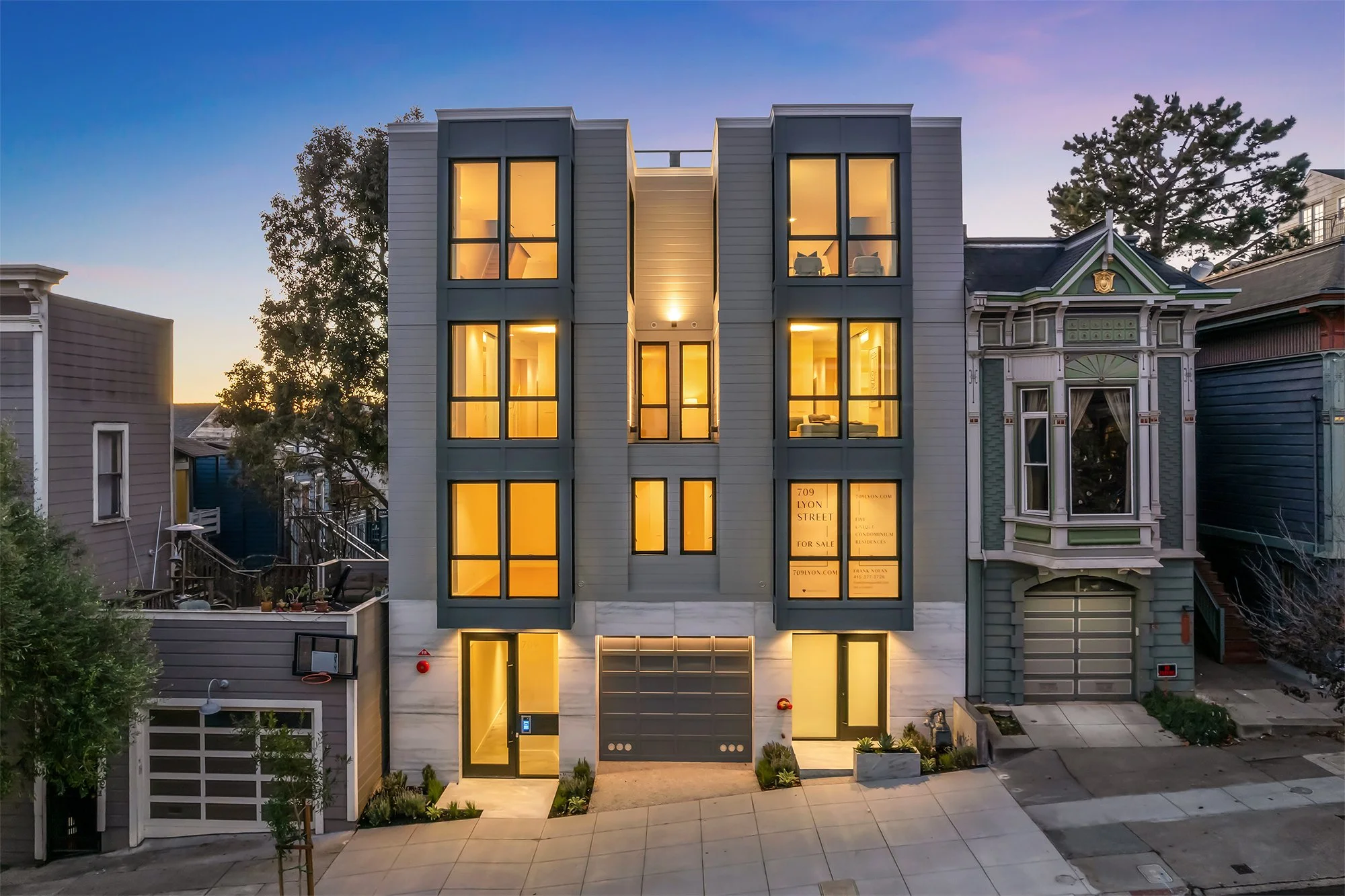 Modern multi-story residential building with large illuminated windows, located in an urban neighborhood during dusk, with neighboring houses and trees visible.