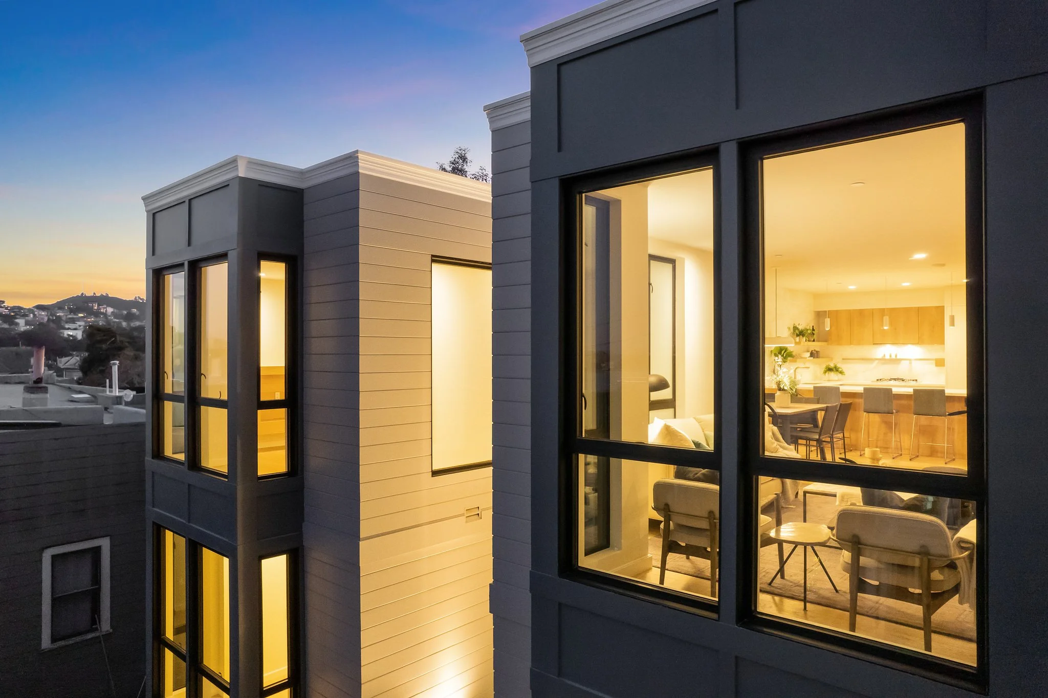 Modern multi-story building with large lit windows overlooking a cityscape at sunset.