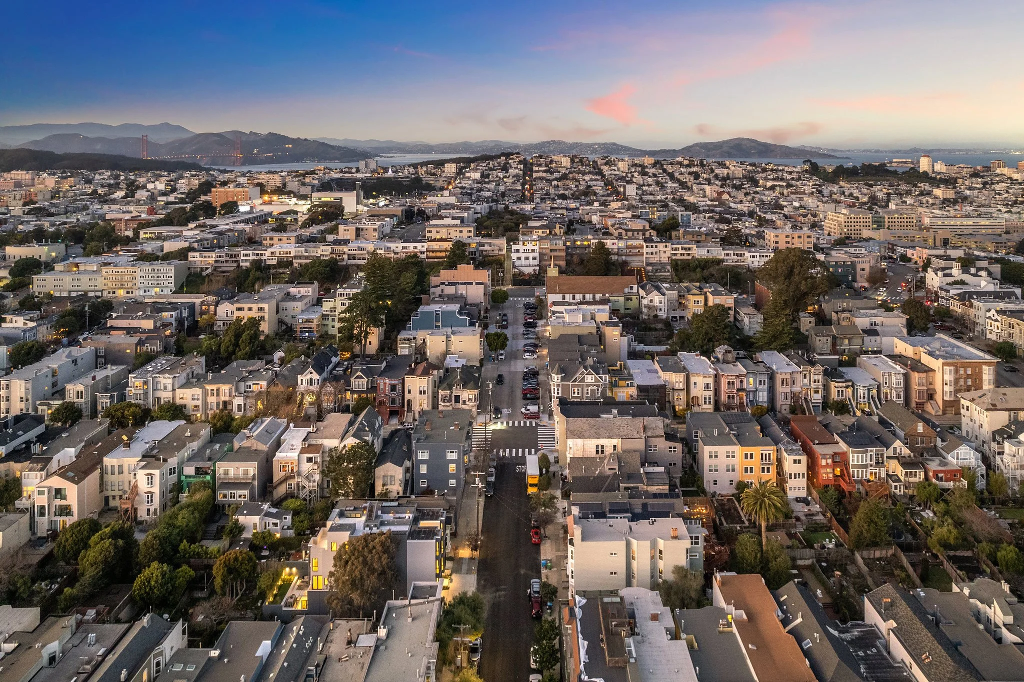A panoramic view of a densely populated urban area with numerous houses and buildings, showing a city skyline with hills and a bridge in the background during dusk.