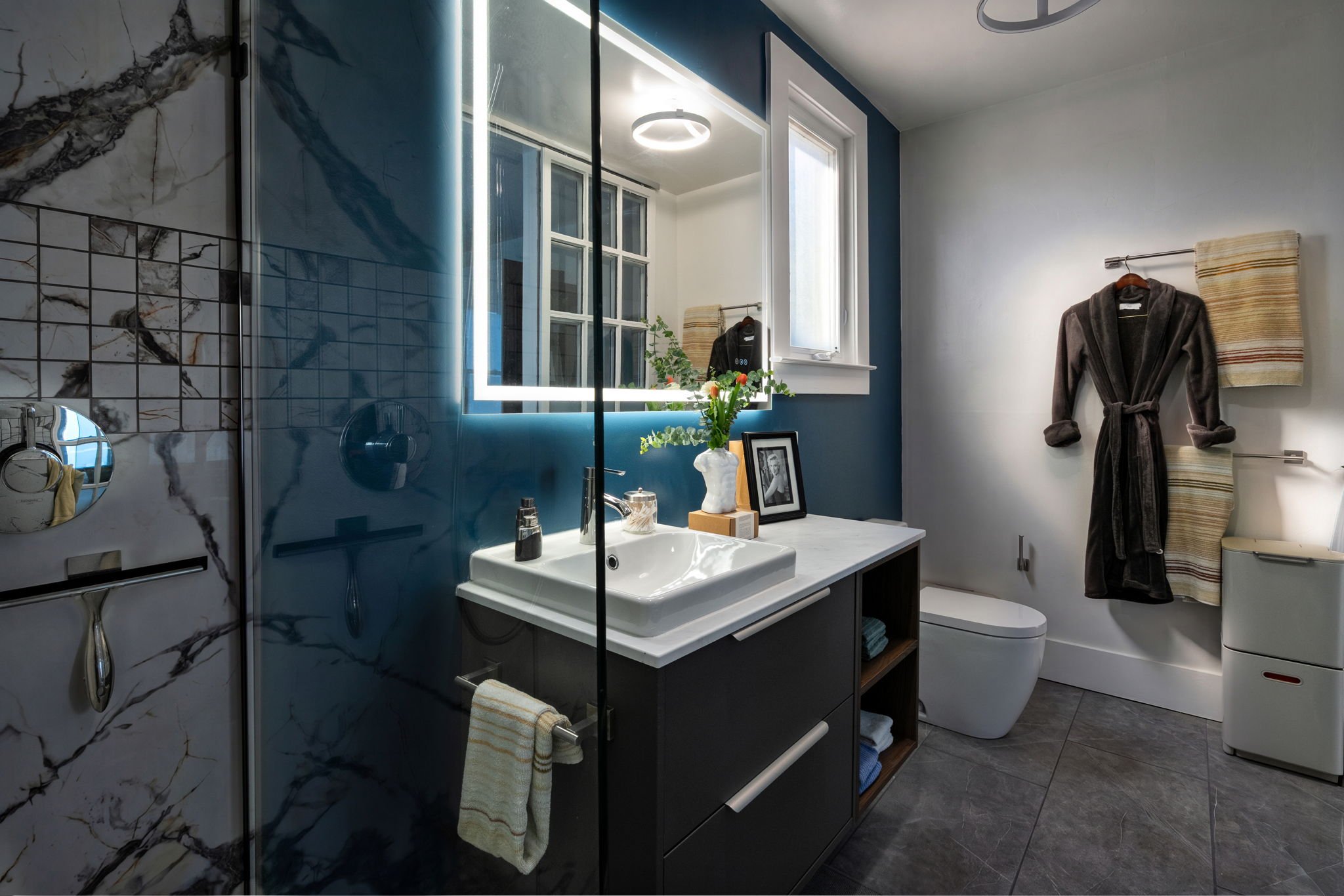 Modern bathroom with marble and tile walls, a mirror with LED lighting, a small vanity with a sink, a window, and hanging towels.