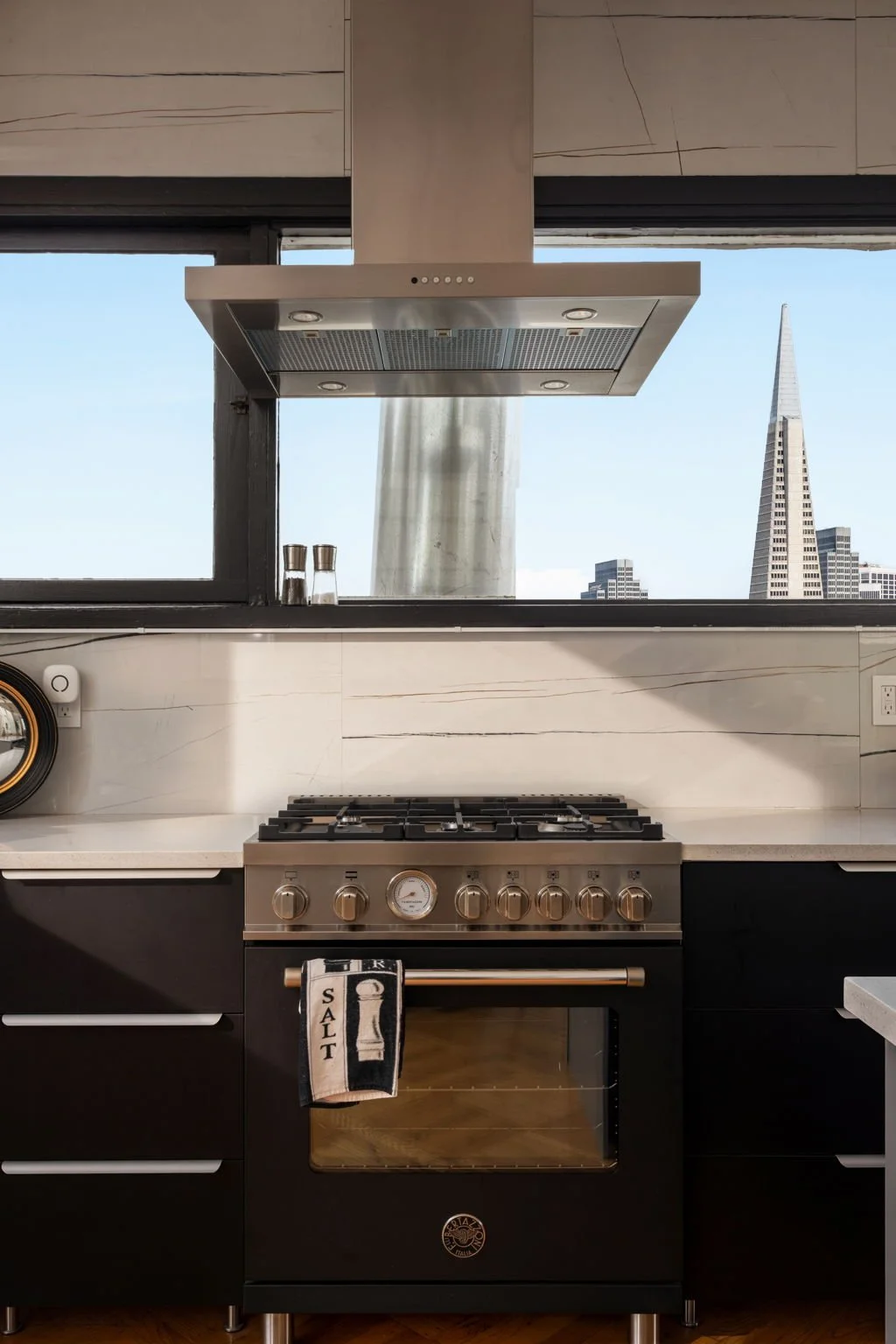 Modern black and silver gas stove with oven, situated in a kitchen with white marble countertops and backsplash. A towel with the word 'Salt' hangs on the oven handle. There is a window above the stove showing the San Francisco skyline, including the