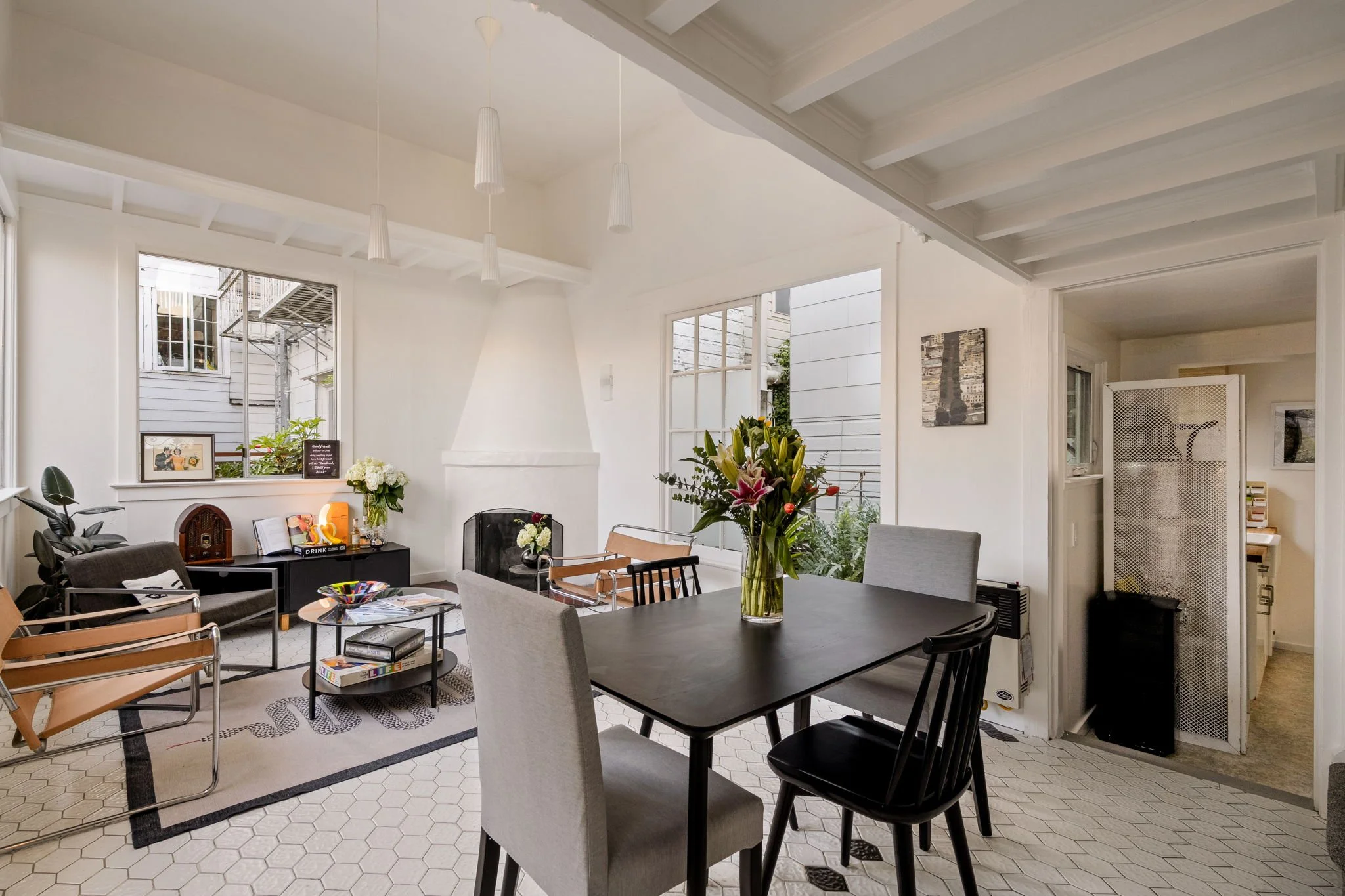 Interior of a bright, modern living and dining room with large windows, white walls, and various furniture including a floral centerpiece, chairs, and a table.