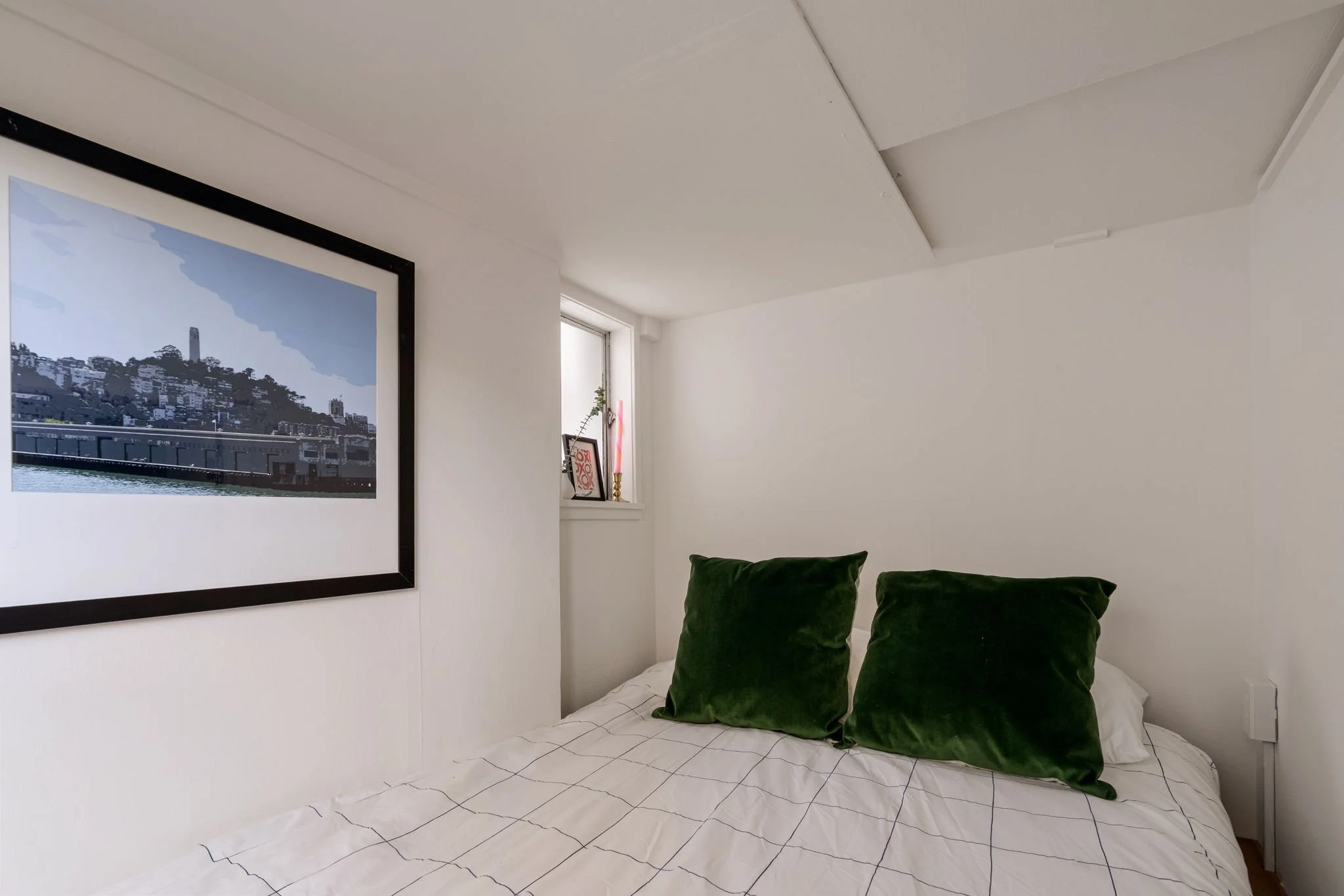 A bedroom with white walls and ceiling. On the wall, there is a large framed picture of a cityscape with a body of water in the foreground. Near the window, there are small decorative items, including a picture frame, a candle, and a small plant. The