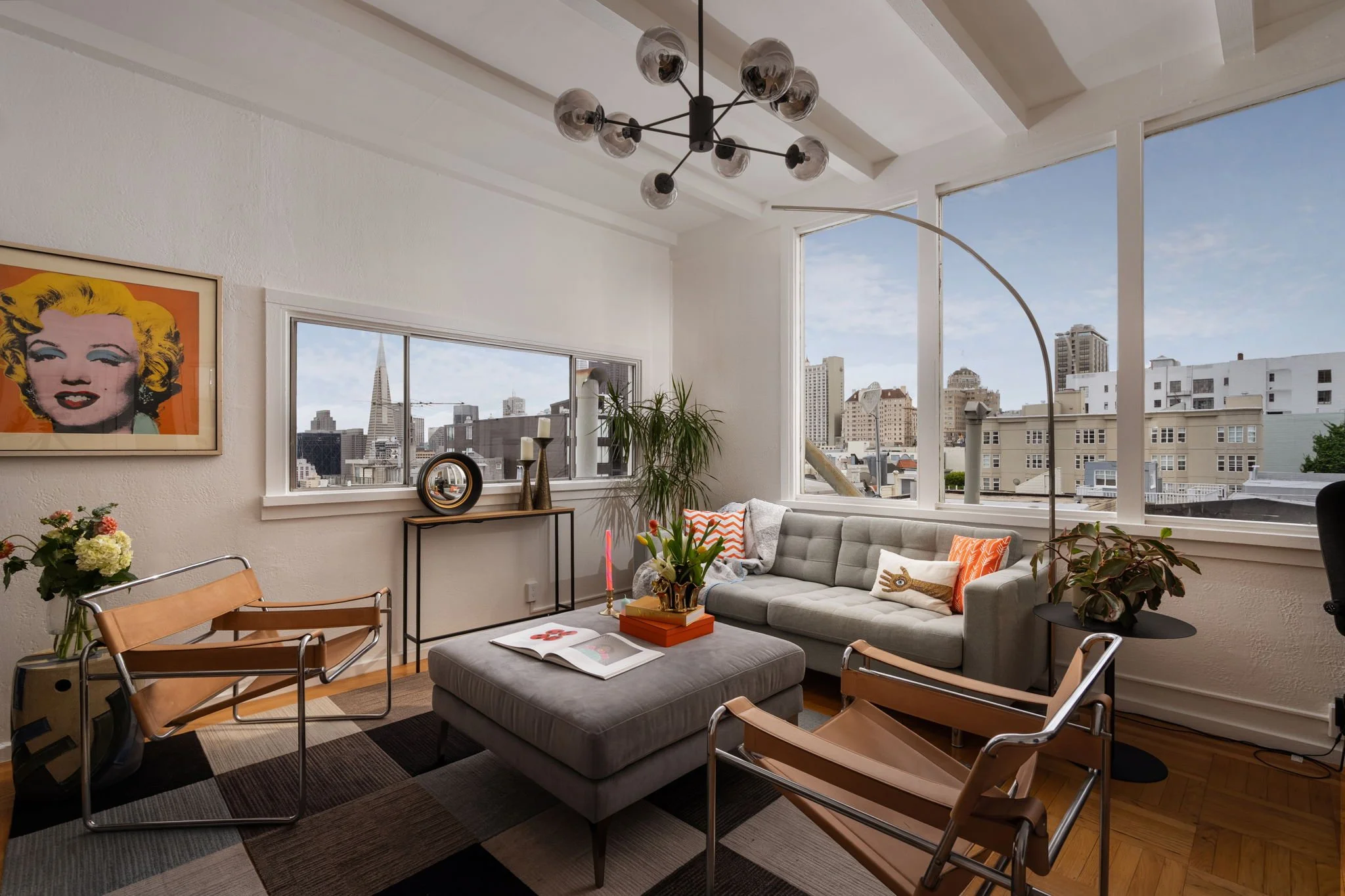 A modern living room with large windows showcasing a city skyline, including the Transamerica Pyramid building. The room has a gray sofa with orange and white pillows, a rectangular ottoman with an open magazine and decorative items, a black side table with candles, and a tall arching floor lamp. There is a colorful abstract artwork on the wall, a potted plant, and a dining area with chairs and a table partially visible in the foreground.