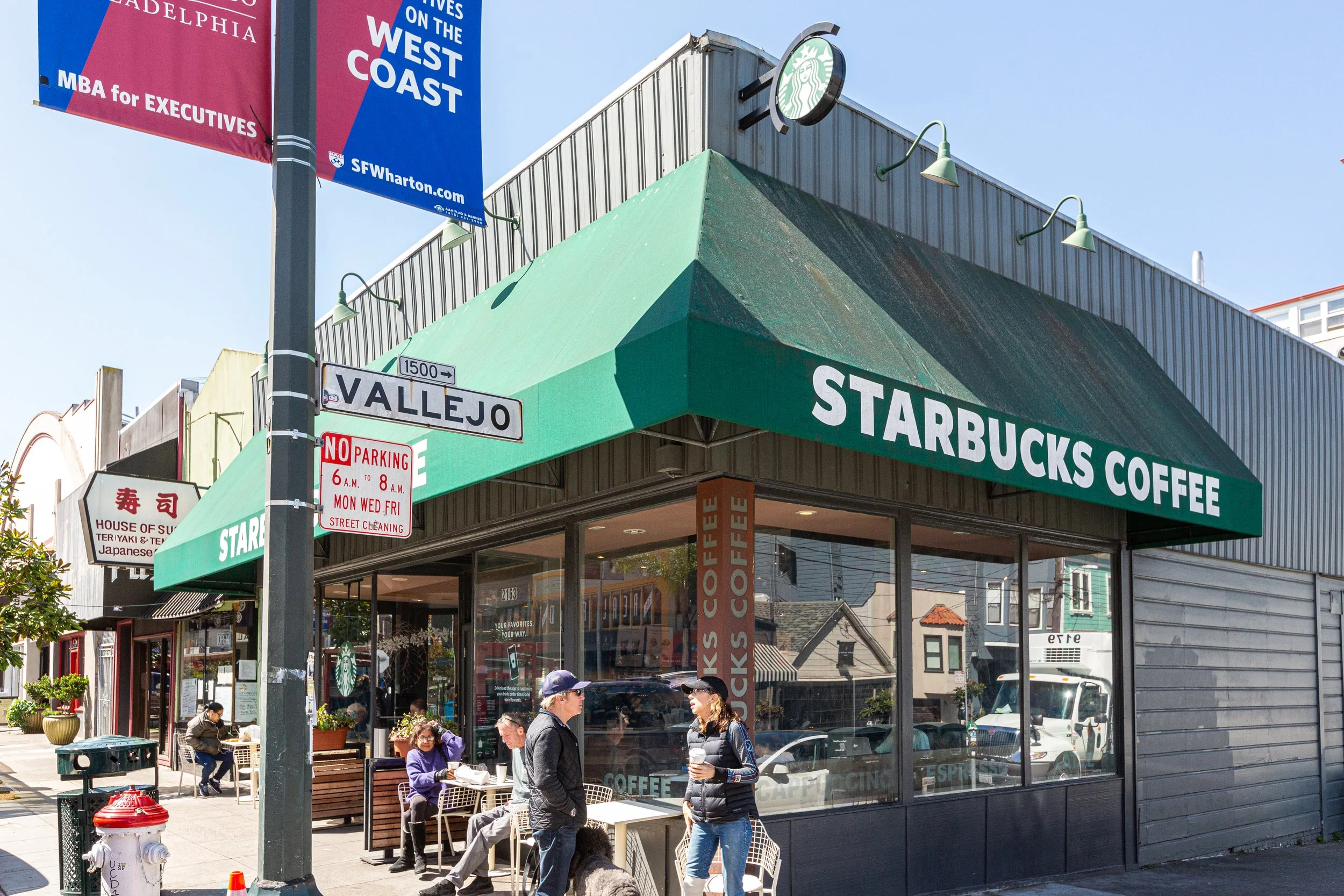 Street view of a Starbucks Coffee shop with an outdoor seating area. Pedestrians are walking and sitting at tables. There are street signs including Vallesjo and no parking signs. Overhead banners advertise an MBA program at SFWharton, and the buildi