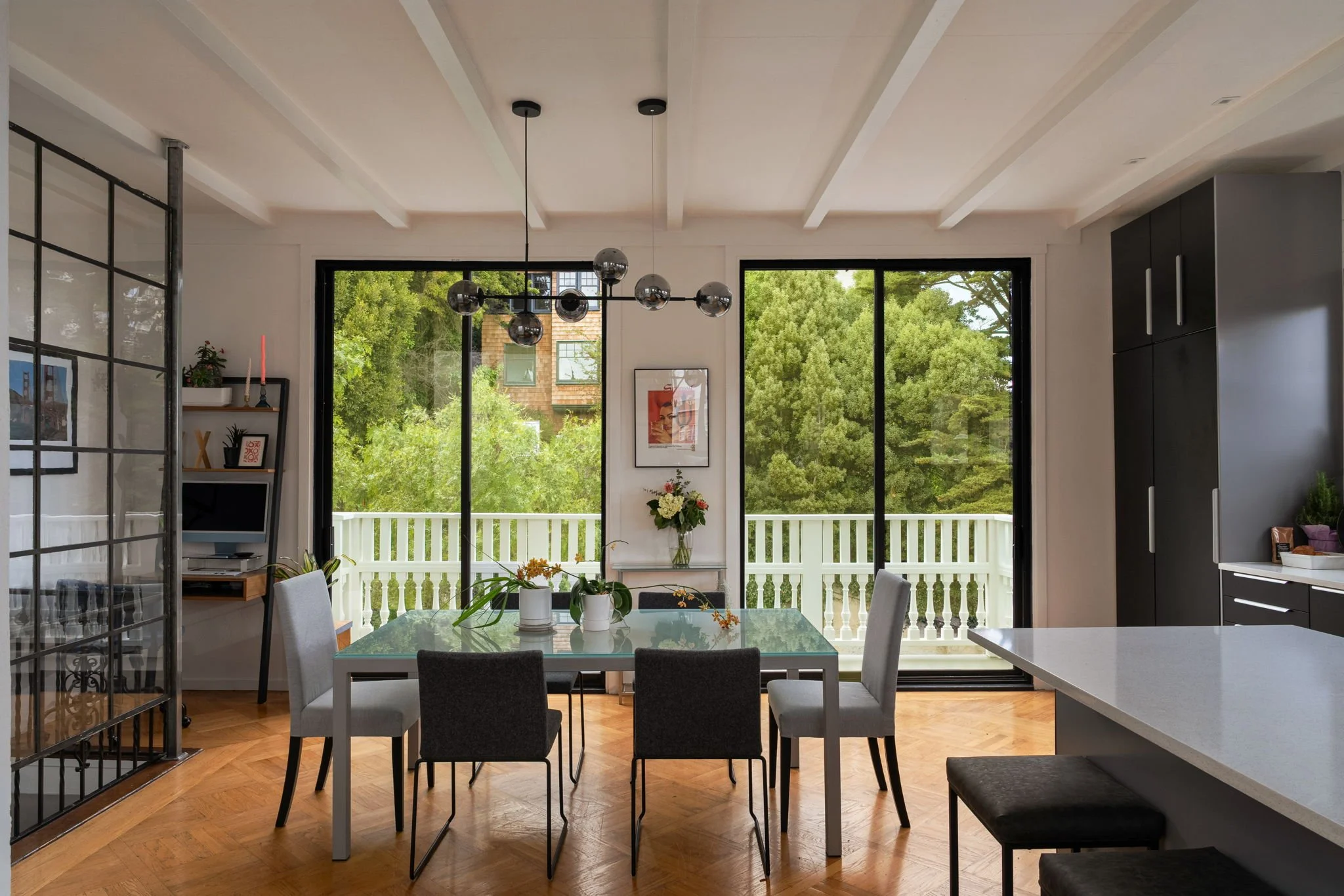 A modern dining area with a glass top table surrounded by eight chairs, a center floral arrangement, large windows with lush green trees outside, pendant lights hanging from the ceiling, and a balcony railing visible through the windows.