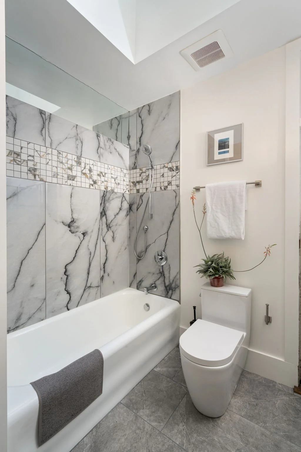 Bathroom with a bathtub and a separate toilet. The bathtub has a marble tile wall with a decorative band of small mosaic tiles, and a handheld showerhead. The toilet has a potted plant on top of the tank, a white towel hanging on a towel bar, and a f