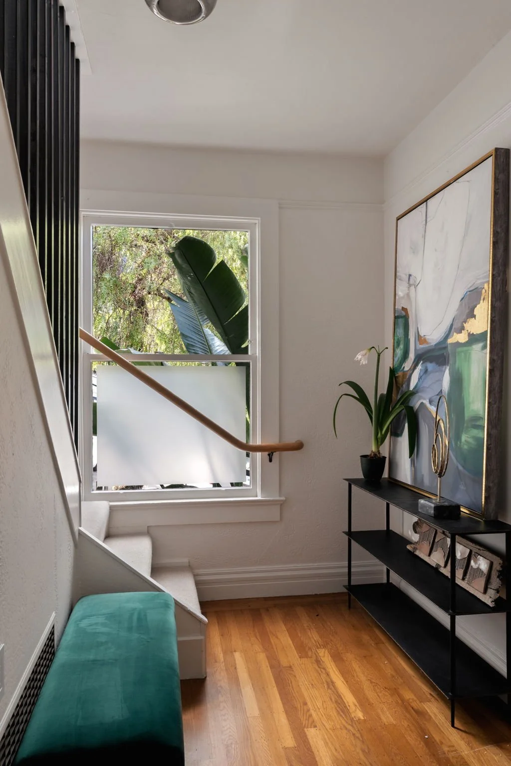 Interior view of a staircase landing with a window showing greenery outside, a teal bench, a black console table with framed pictures and decorative items, a large abstract painting, a potted plant, and a staircase with a wooden handrail.