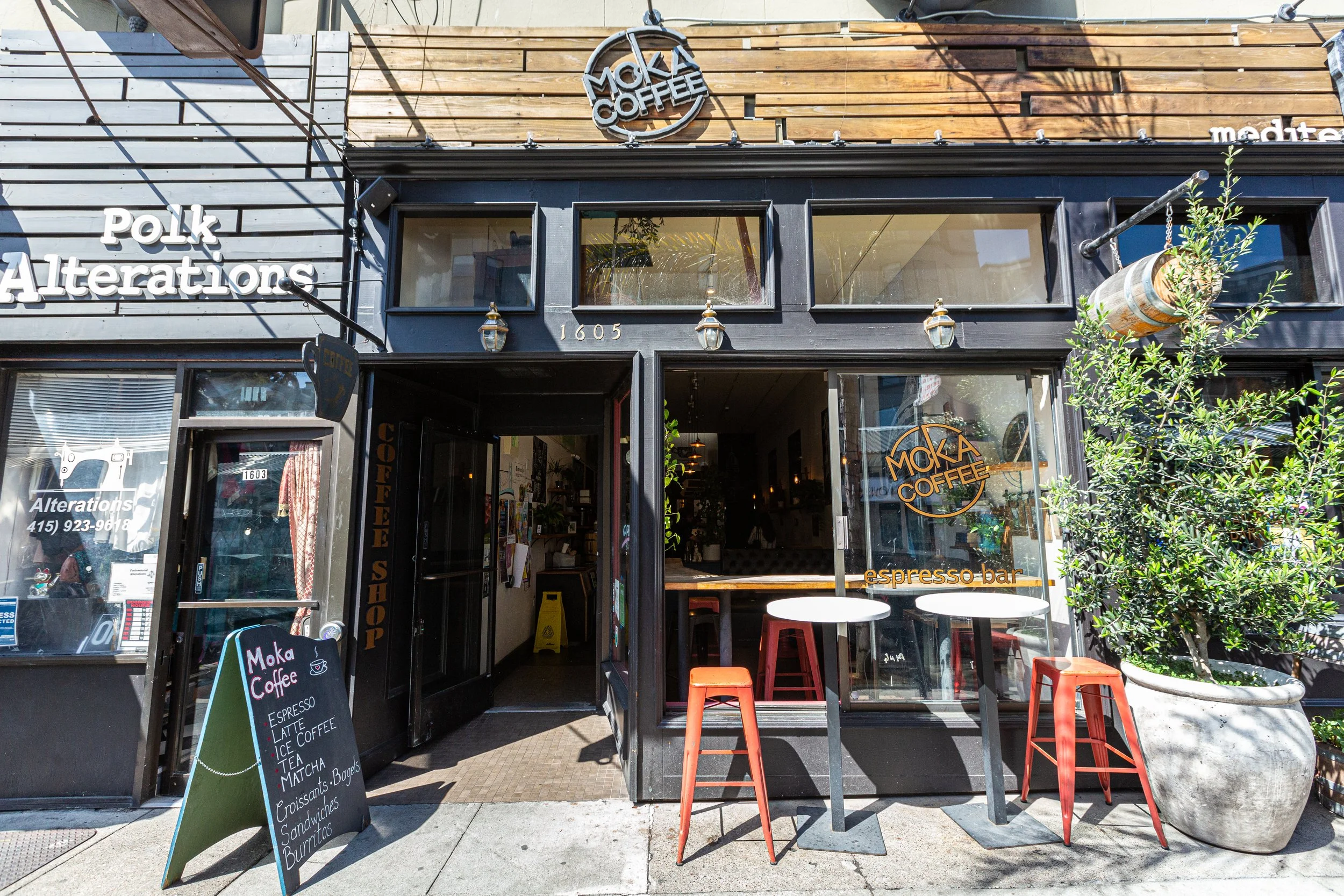 Exterior of Moka Coffee shop with black framing, wooden accents, and outdoor seating featuring three red metal stools and a small round table. Signage includes the shop's name and menu offerings for espresso, latte, ice coffee, tea, matcha, bagels, c