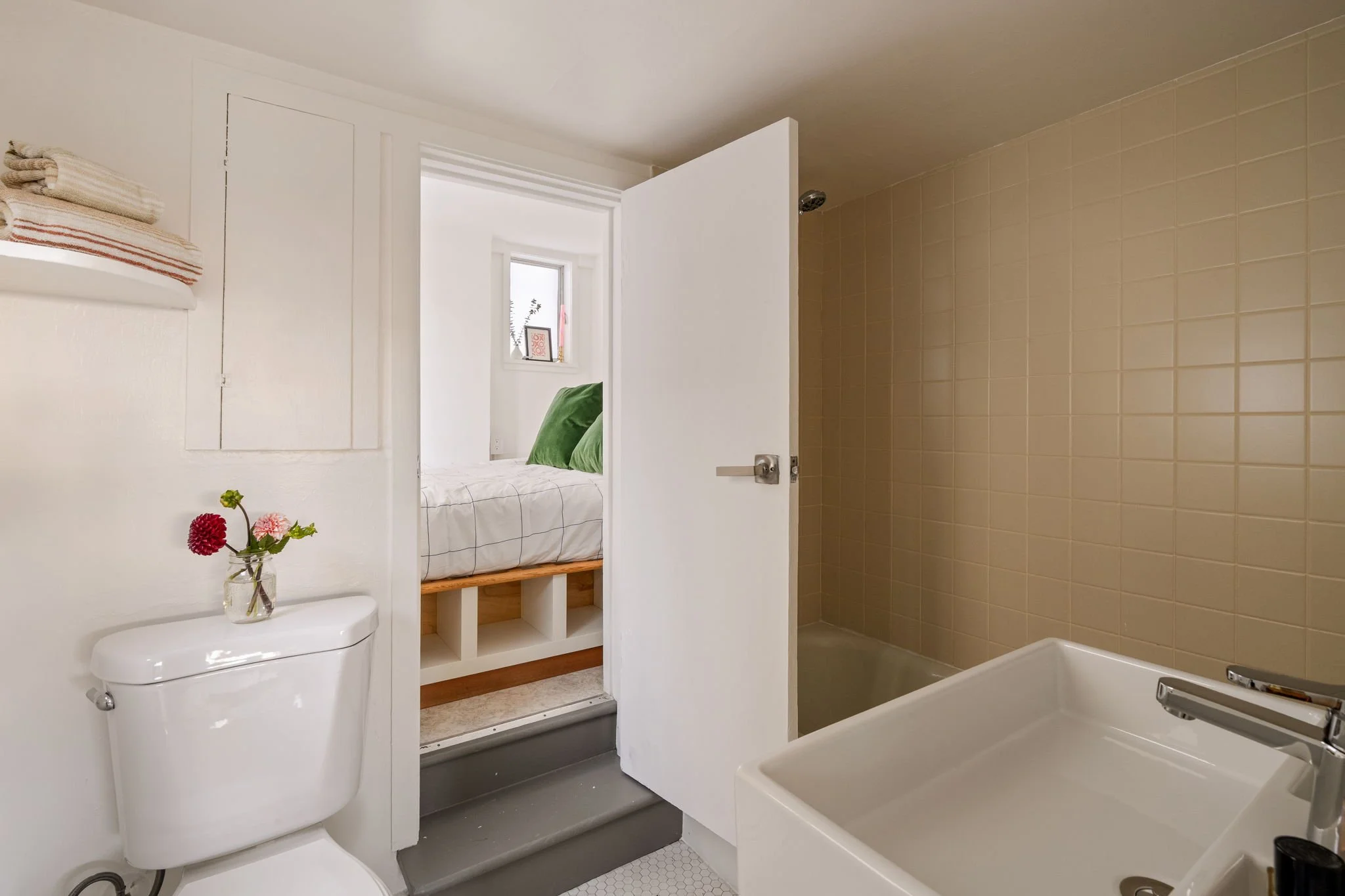 A small bathroom with a white toilet, a small vase of flowers on top, a white shelf with folded towels, a bathtub with a faucet, and a shower area with beige tiles. A bed with green pillows is visible through a partially open door on the other side.