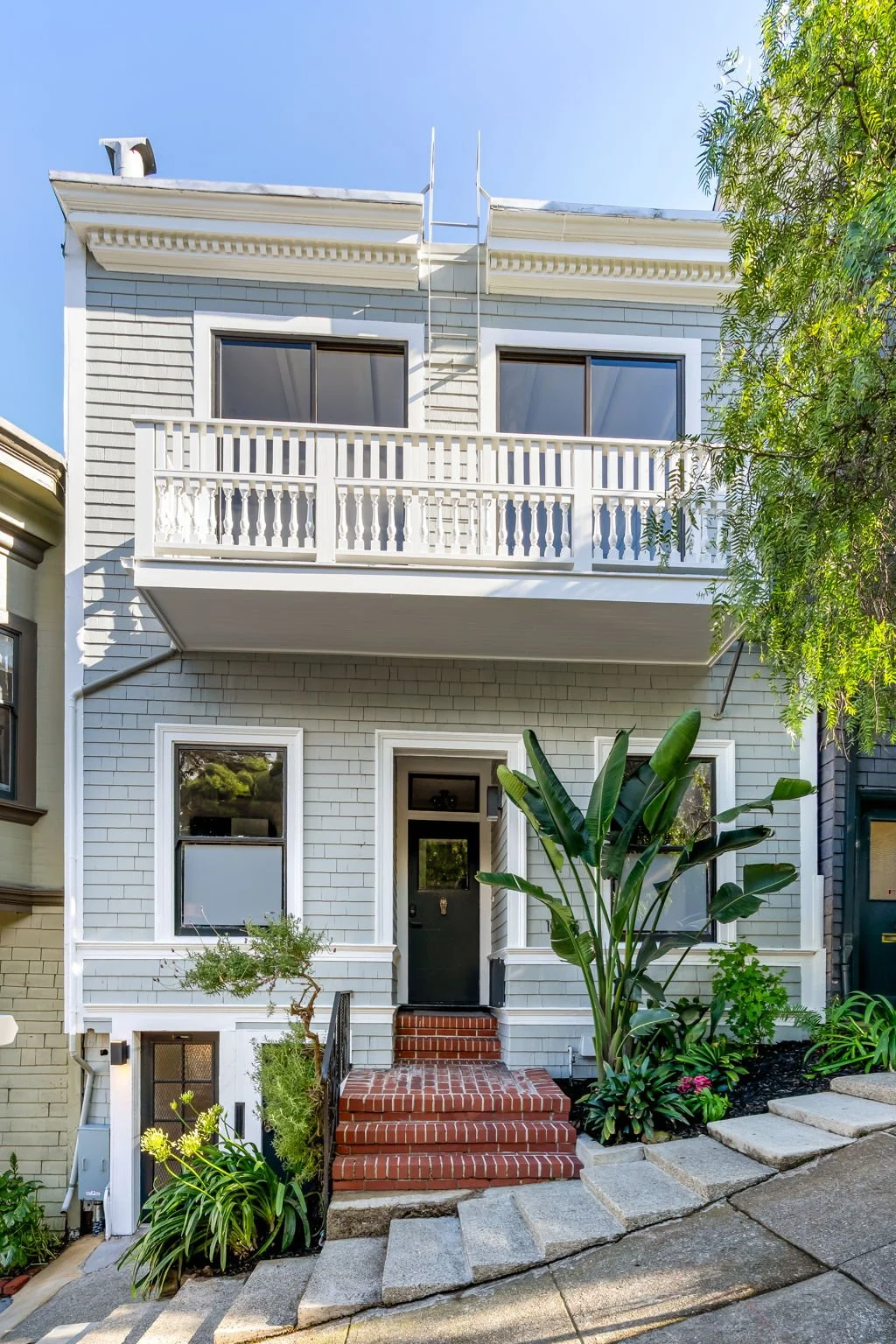 A three-story house with light gray shingle siding, black front door, brick steps, small garden with plants, and a balcony on the second floor with white railing. Clear blue sky above.