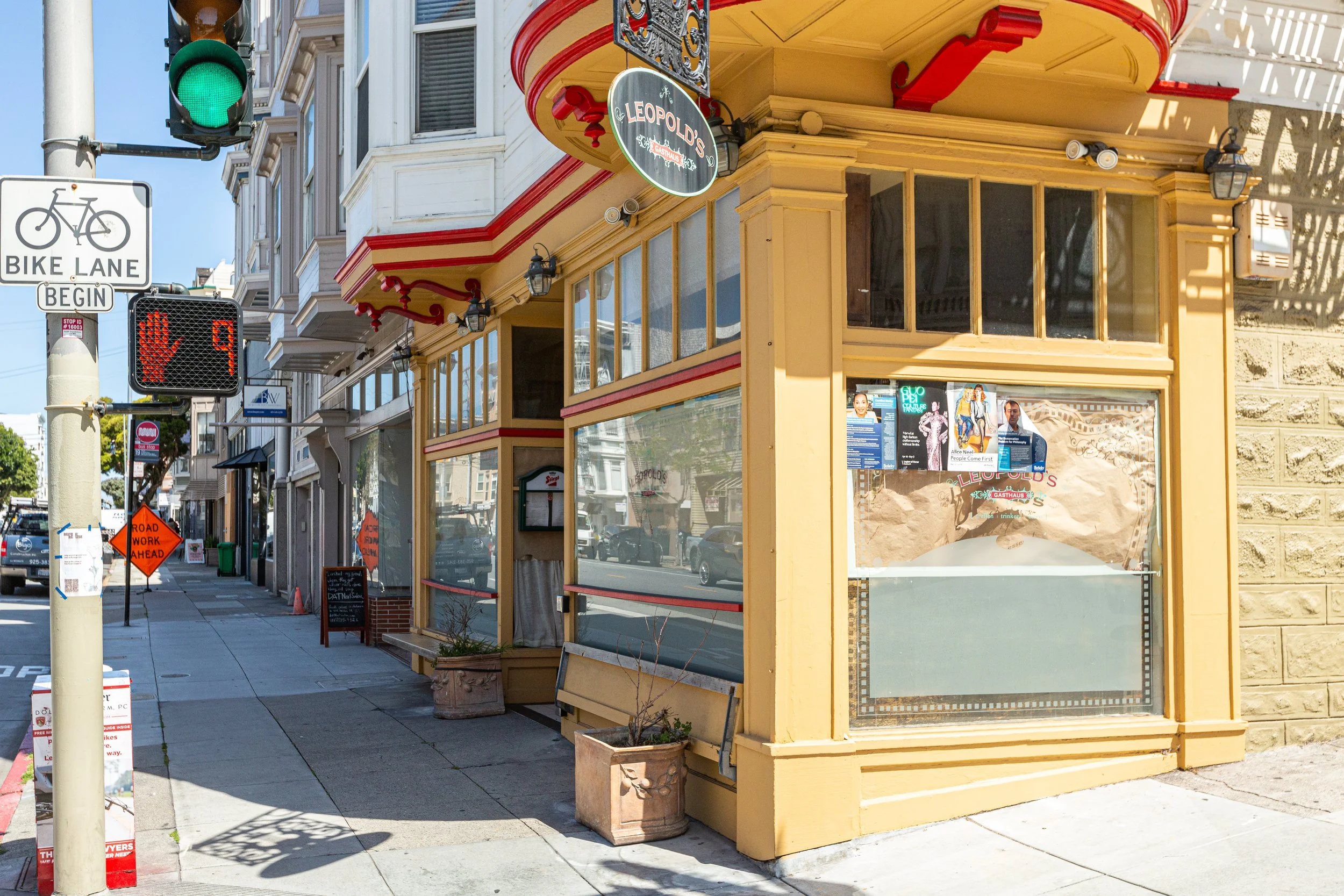 Street scene showing a traffic light, a bike lane sign, and a corner storefront with yellow and red trim that reads 'Leopold's.' The storefront has large display windows with posters and a sign hanging above. There are sidewalk signs and a constructi