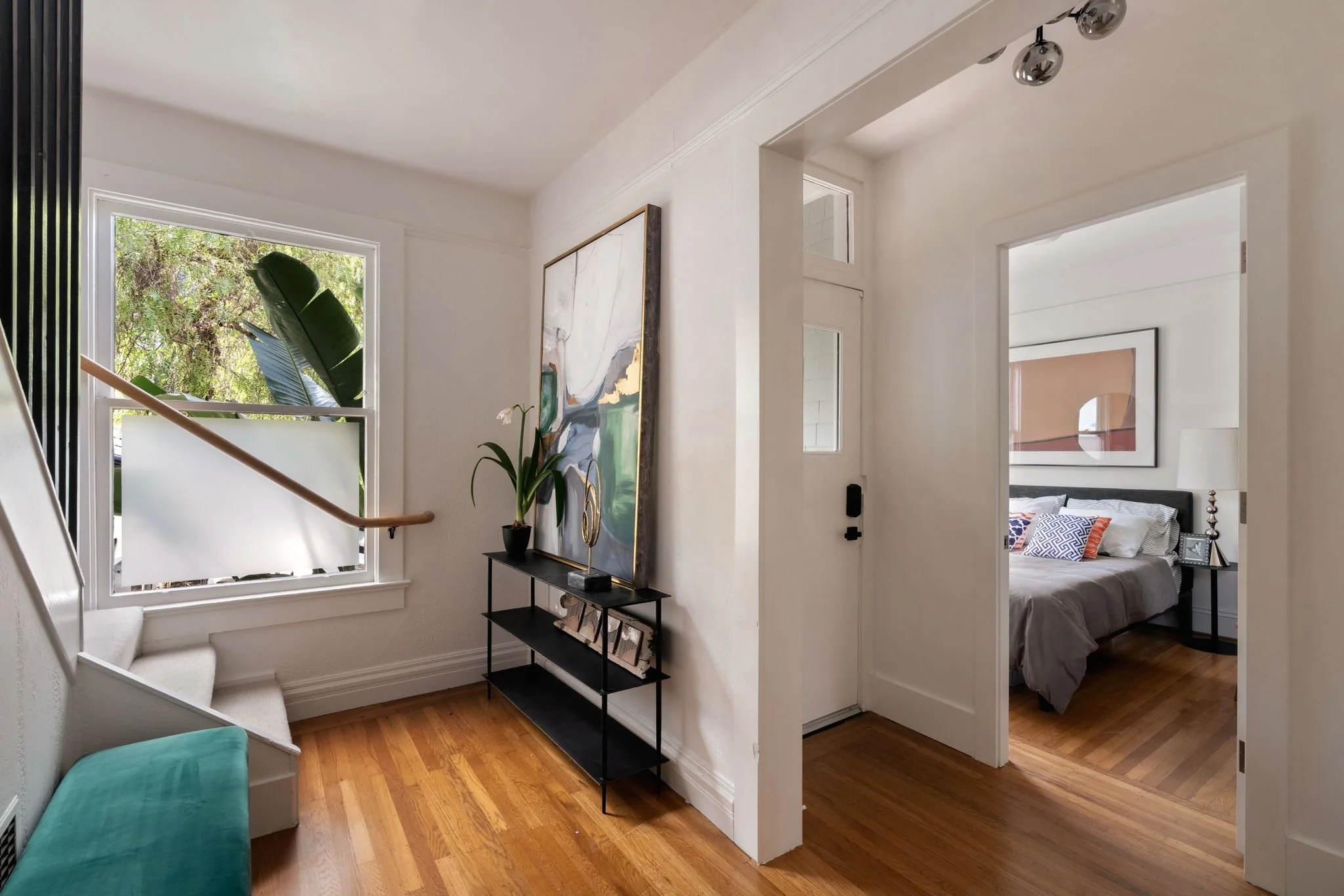 A staircase with a window overlooking green trees, a side table with decor and a large painting, and an open door leading to a bedroom with a bed with patterned pillows.