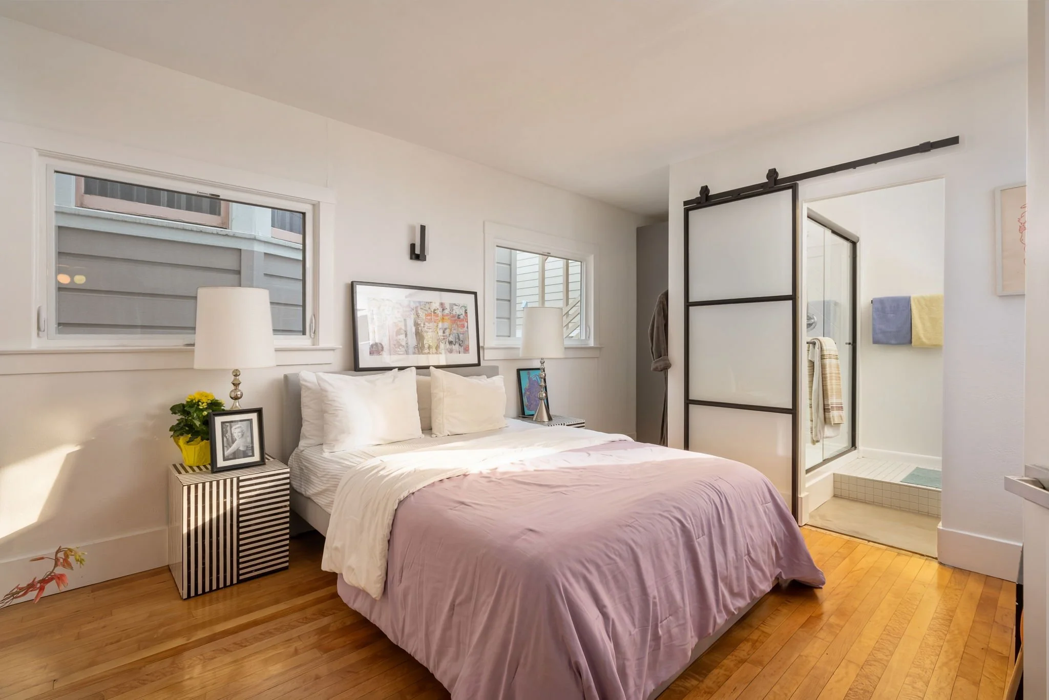 A cozy bedroom featuring a bed with white pillows and a pink bedspread, side tables with lamps, framed artwork, and large windows with view of neighboring houses. An adjacent bathroom with towels and a shower is visible.