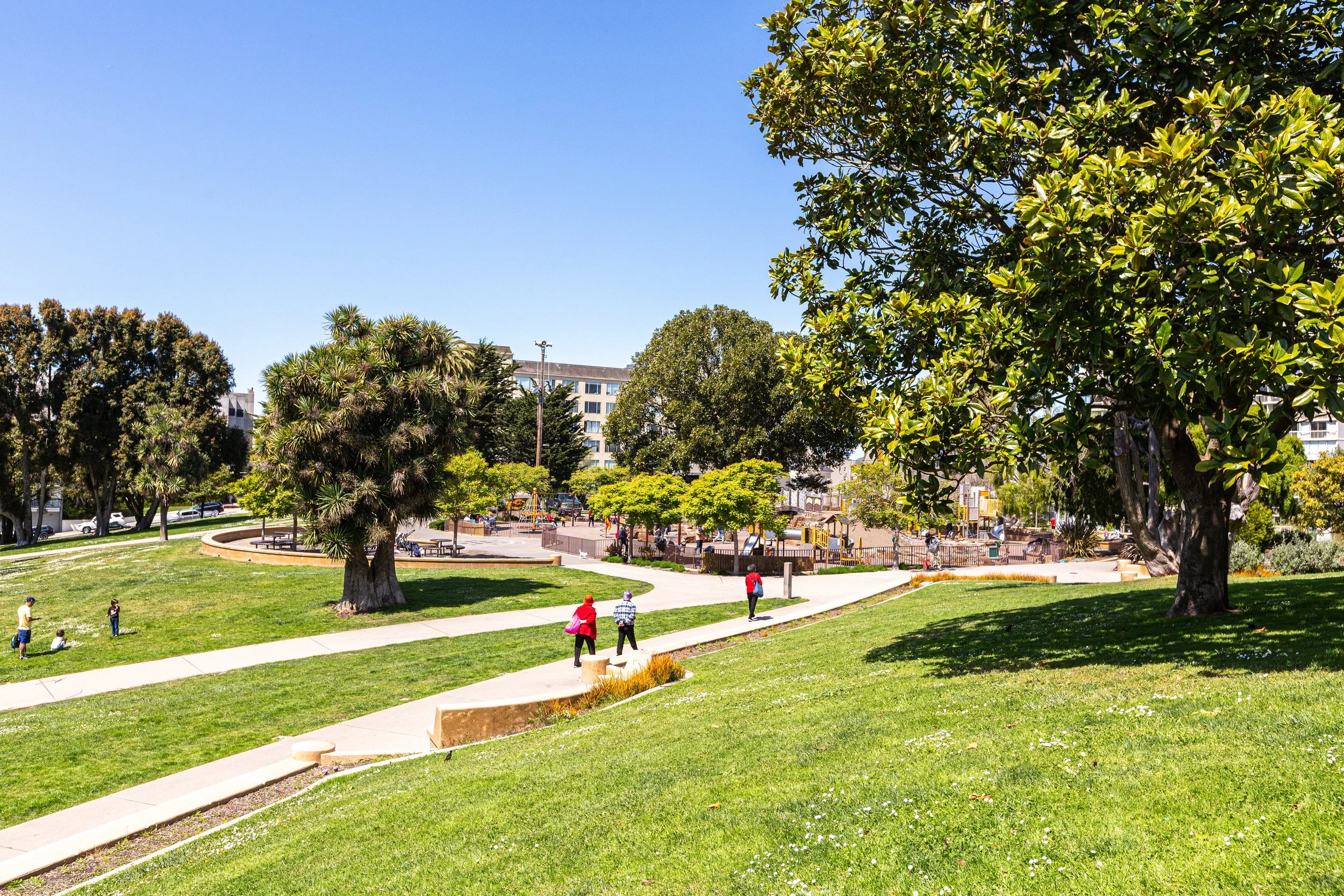 A park with lush green grass, winding concrete paths, and various trees. There are people walking, including children playing and adults strolling. In the background, there is a playground area with yellow and purple equipment, and multi-story buildi