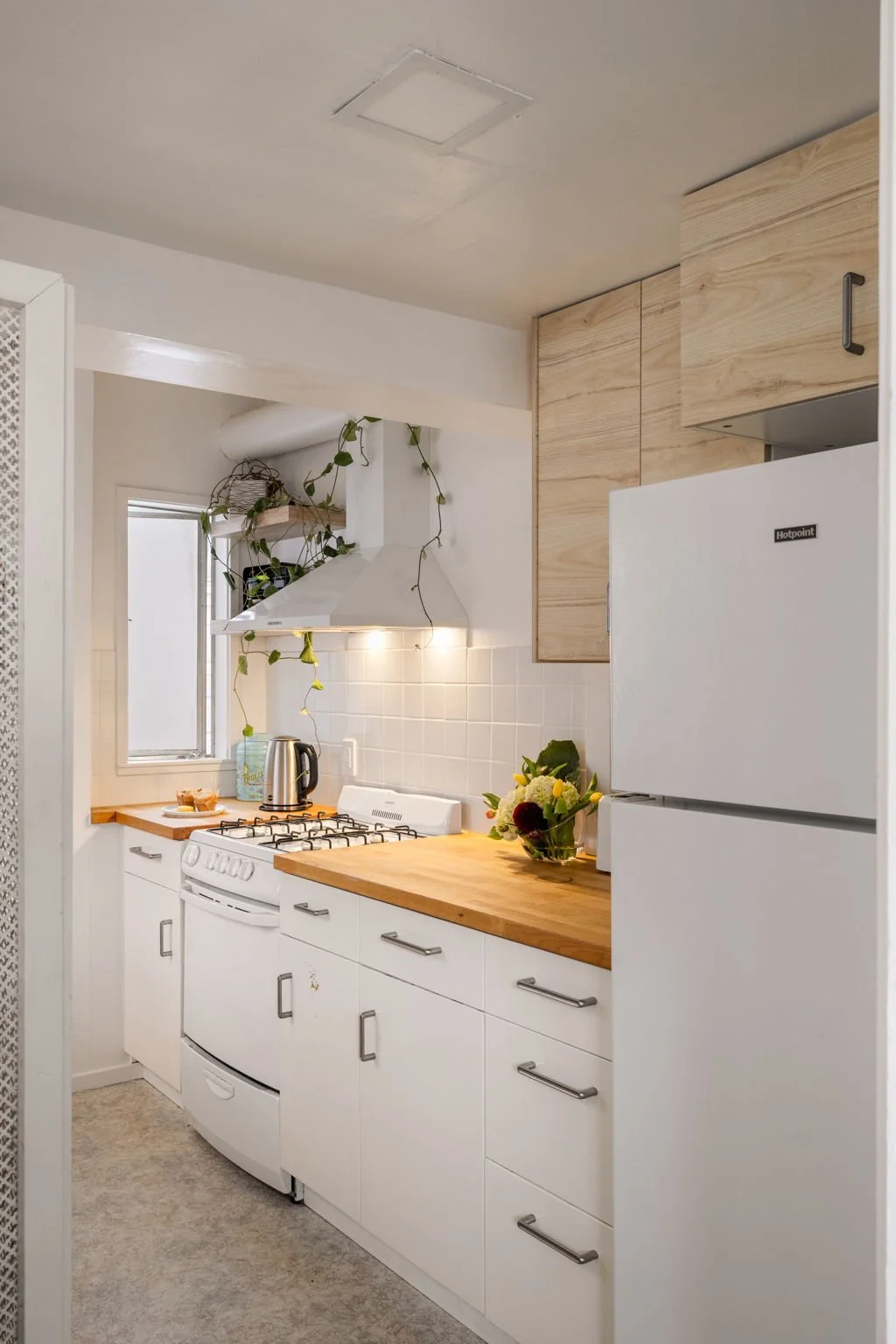 A cozy kitchen with white cabinets, a wooden countertop, a white refrigerator, and a gas stove. There is a small window, hanging plants, a vase with flowers, and a kettle on the counter.