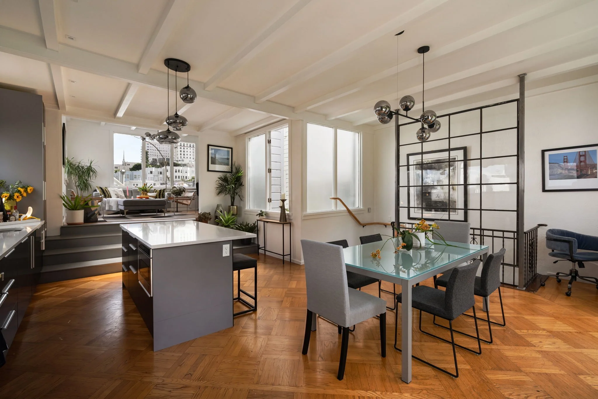A modern open-concept living and dining area with a kitchen island, gray and white furniture, large windows, potted plants, wooden floor, and framed photos on white walls.