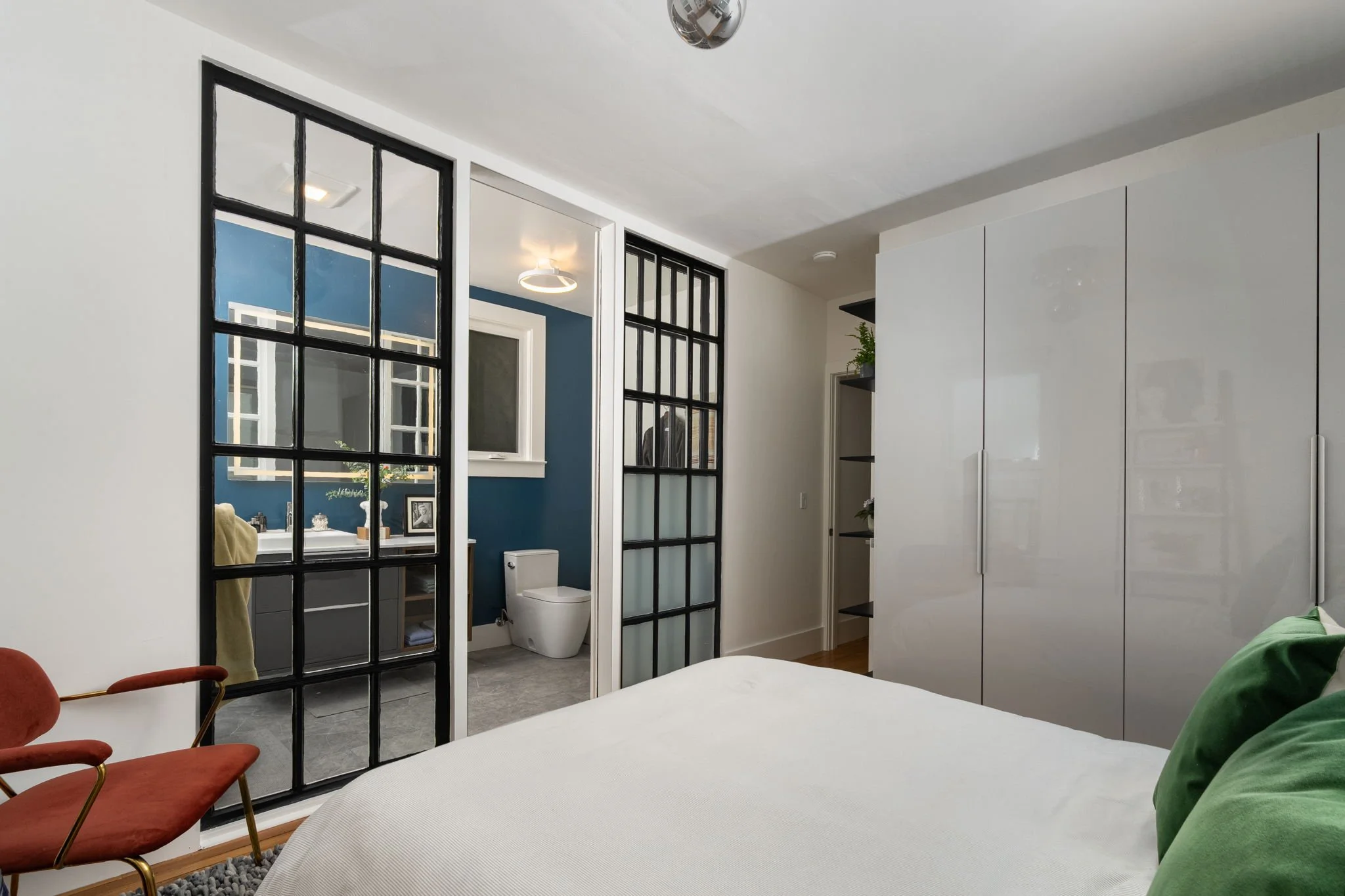 A bedroom with a white bed, green pillow, and two red chairs near a black-framed glass partition that separates the sleeping area from an en-suite bathroom with a toilet. The bathroom has a blue accent wall, and there is a window reflecting outside. 