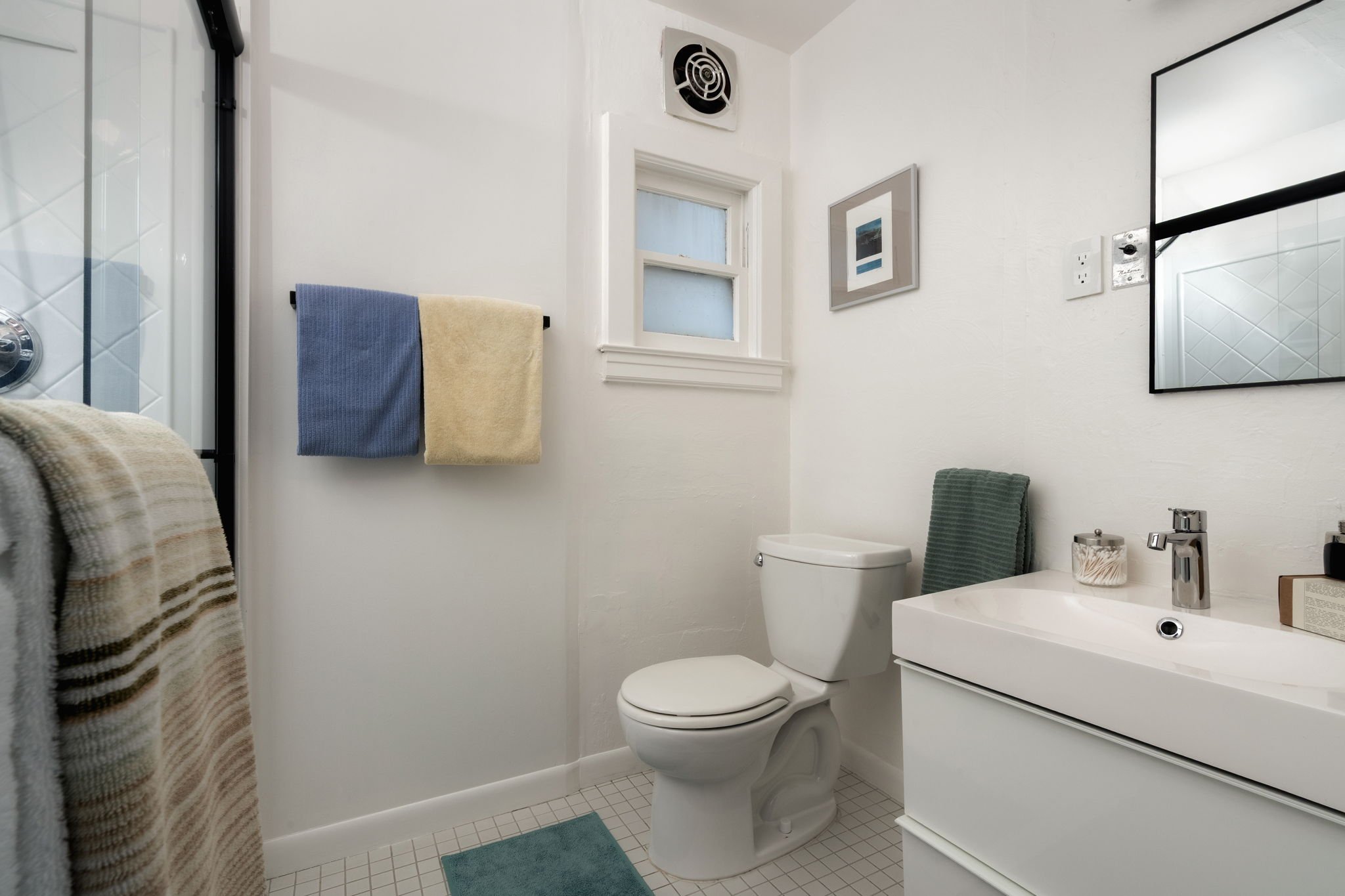 A small white bathroom with a toilet, a white vanity, a mirror, and a small window. There are towels hanging on a towel rack and a teal bath mat on the tile floor.