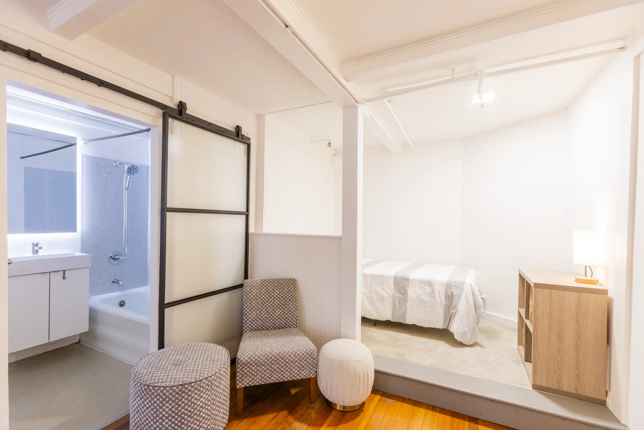 Interior of a small room with a bathroom area on the left, featuring a shower and sink, and a sleeping area on the right with a bed covered in white sheets, a wooden side table, and a patterned upholstered chair and ottomans. The room has white walls