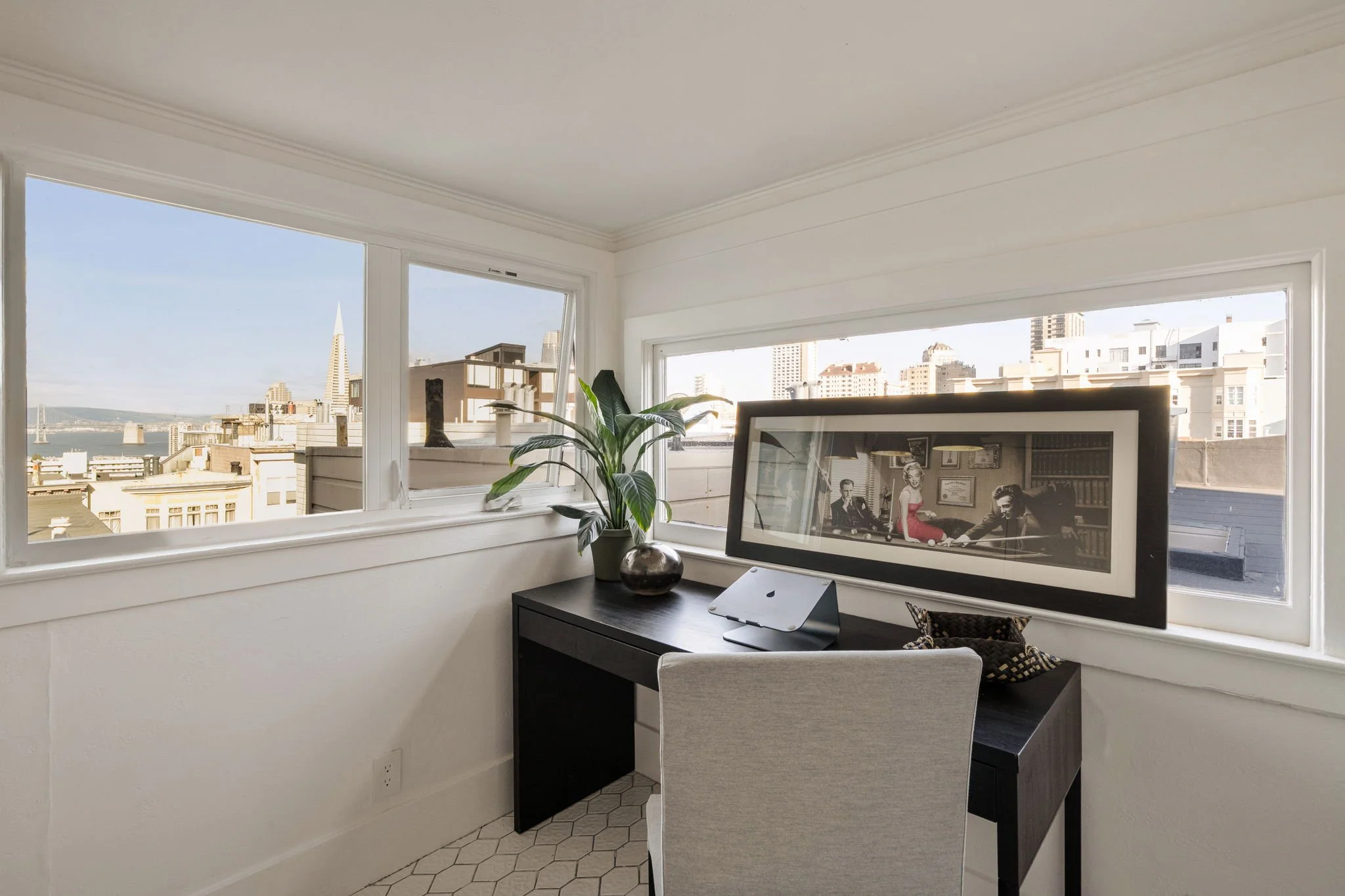 Room with large city view windows, a black desk with a framed black and white photograph, a potted plant, and a chair on a hexagonal tile floor.