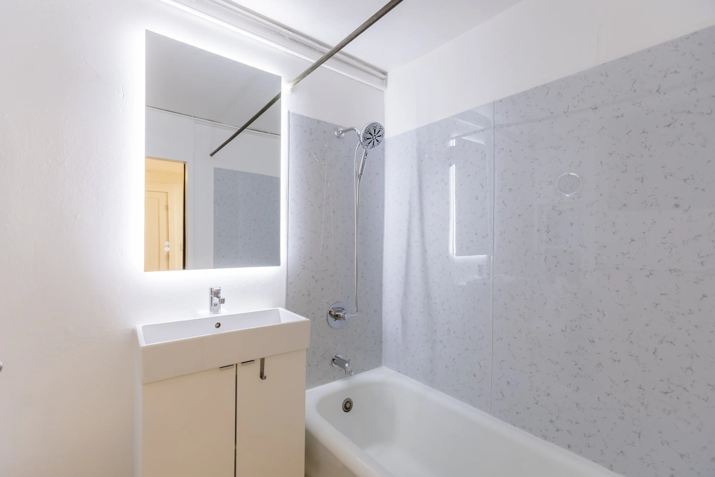 A modern bathroom with a white sink, a mirror with LED lighting, a showerhead, and a tub, featuring white walls and minimalist fixtures.