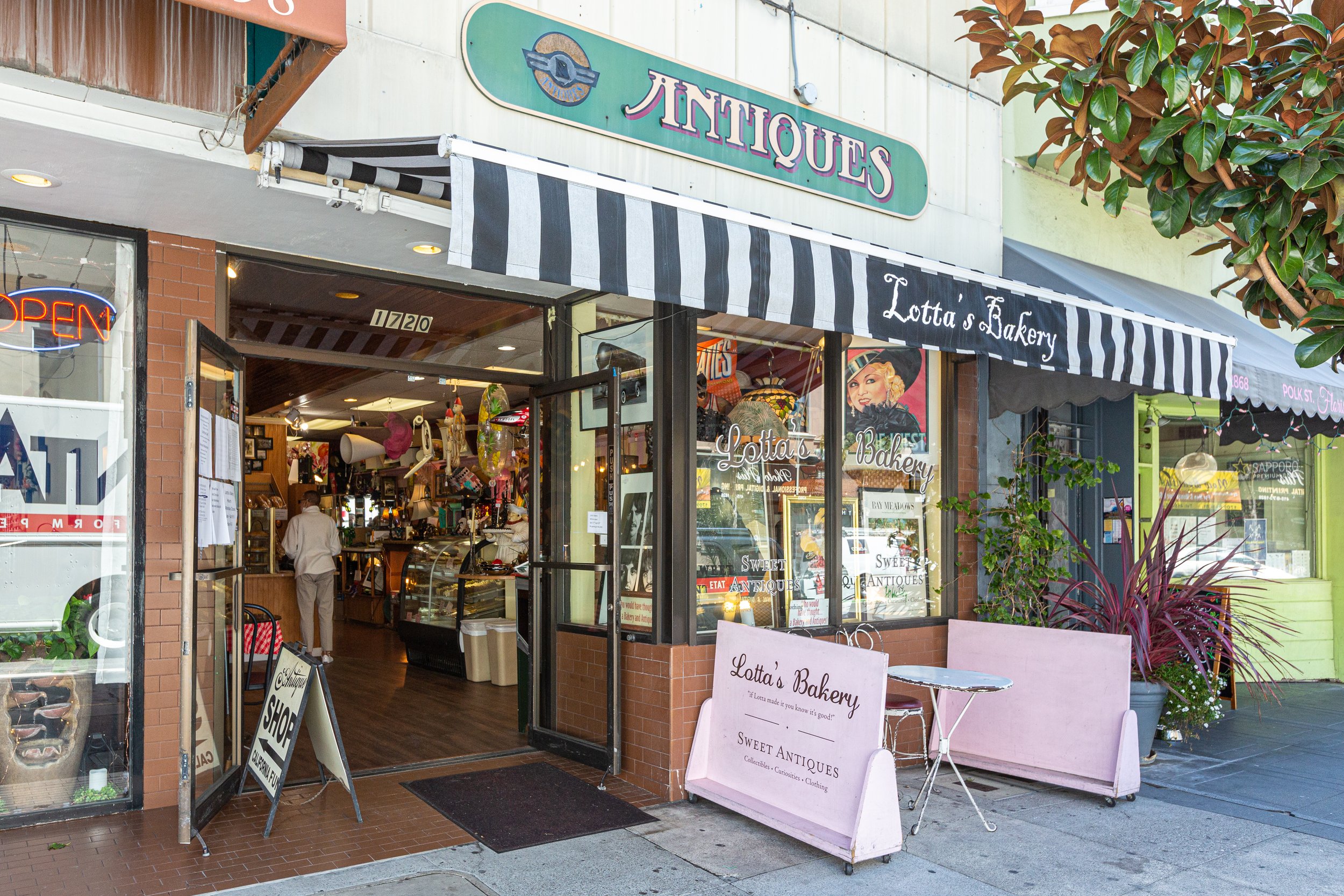 View of a boutique shop called 'Lotta's Bakery' with a partially open door, colorful sign, and striped black-and-white awning. The storefront has large glass windows displaying items inside, with sidewalk signs outside advertising antiques, collectib
