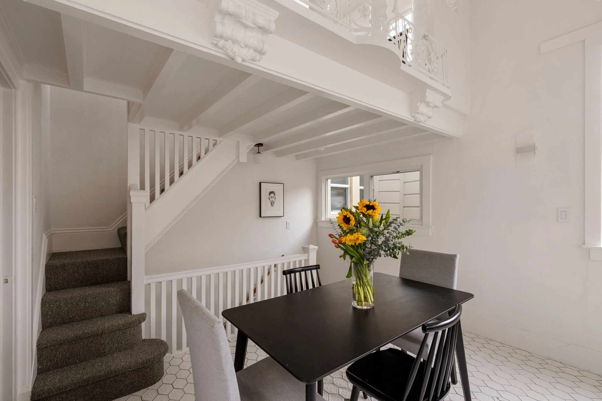 A modern dining area with a black table and black chairs, a clear glass vase of sunflowers on the table, a window with white blinds, white walls, a staircase with gray carpet, and a framed black-and-white drawing on the wall.
