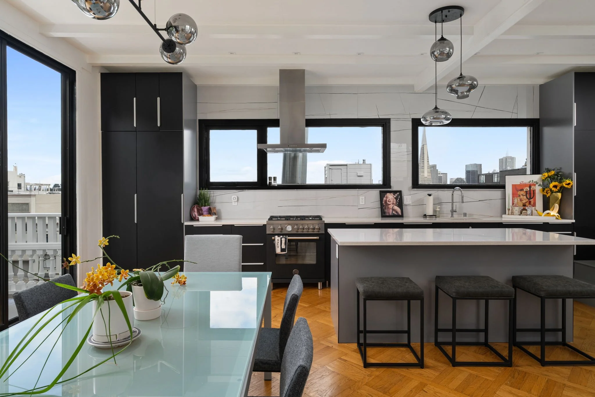 Modern kitchen with black cabinets, white countertops, large windows showing city skyline, and a dining table with potted orchids.