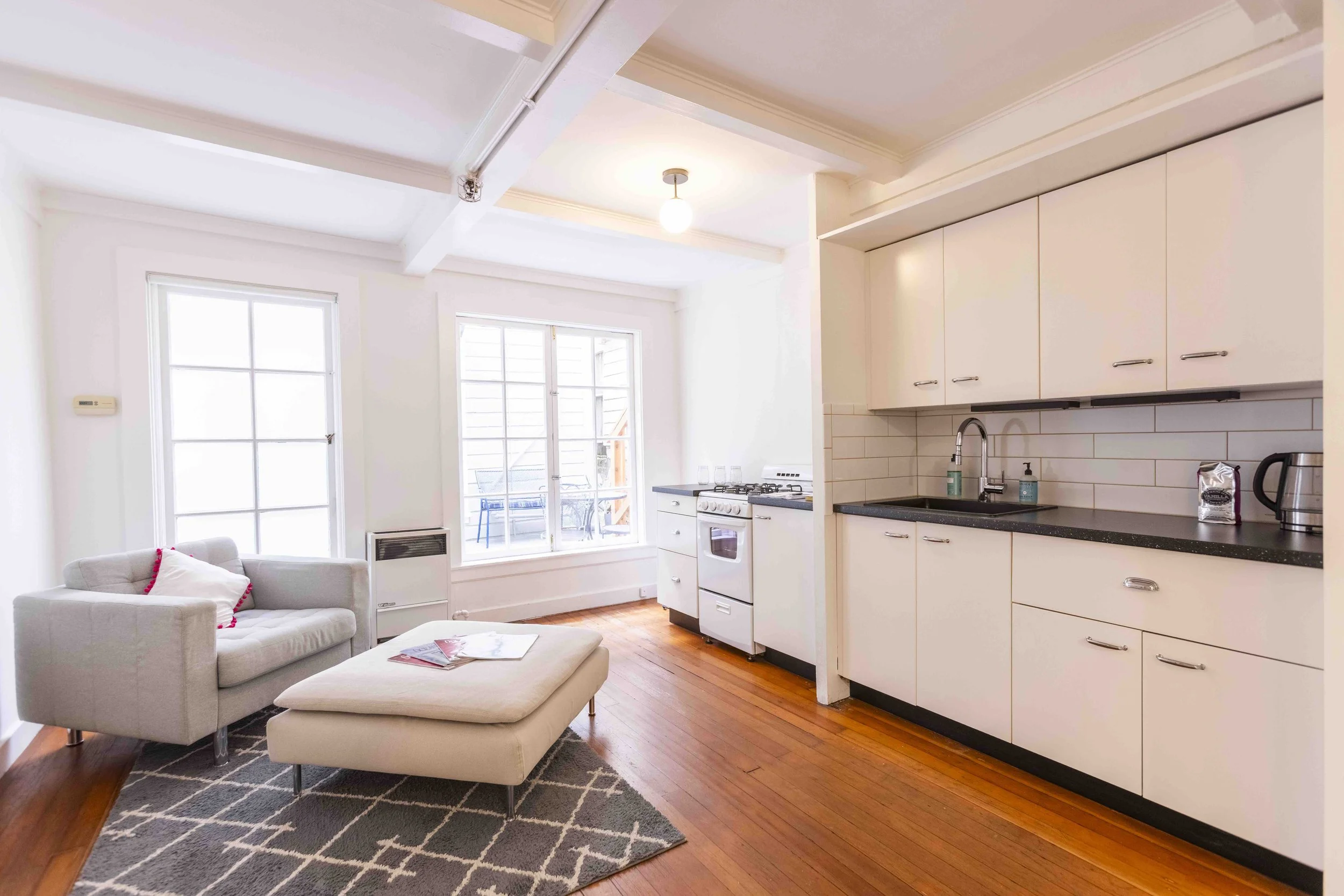 Open-concept living room and kitchen with white walls and cabinets, hardwood floors, large window, and sliding glass door leading to a balcony with outdoor chairs.