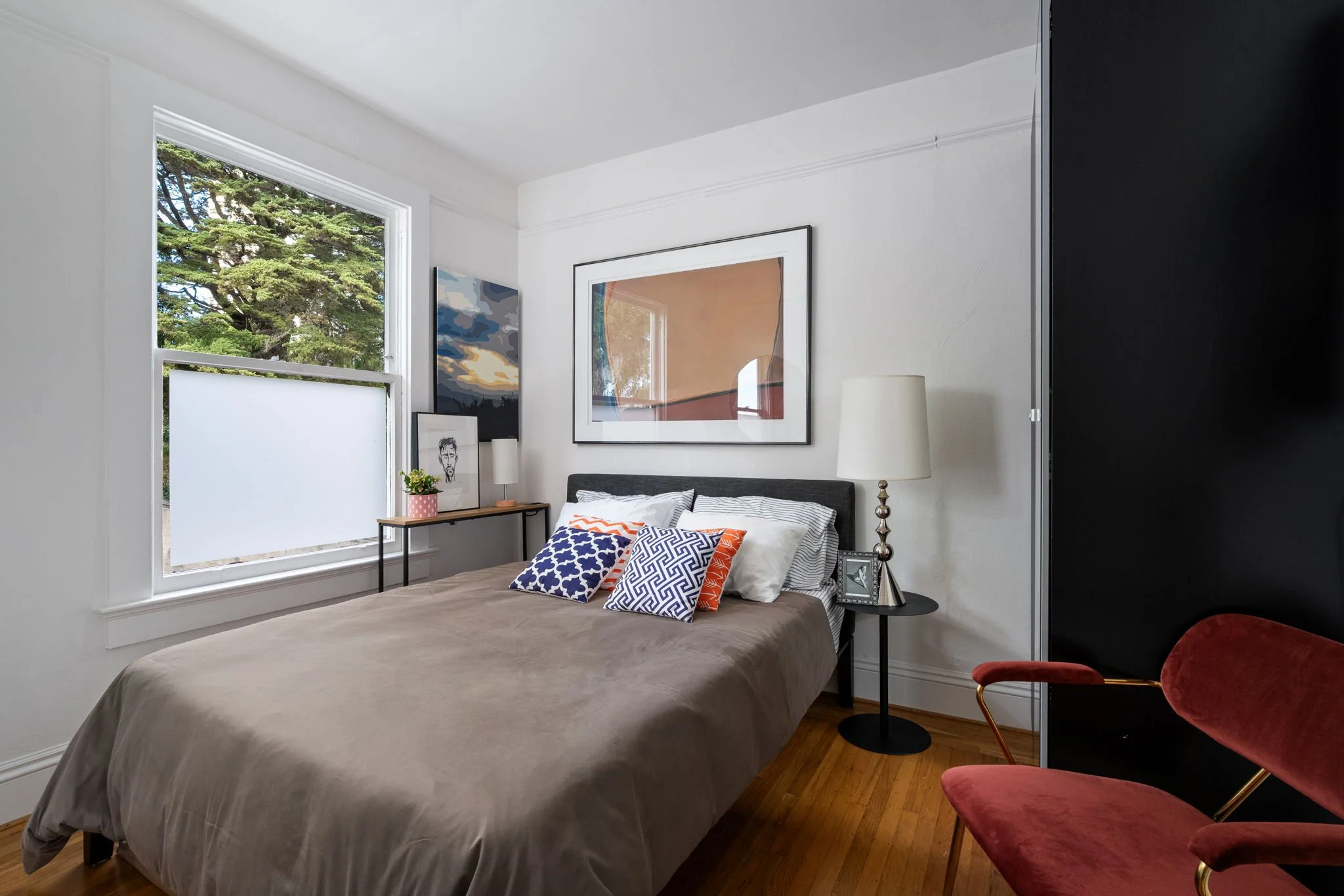 A cozy bedroom with a large window showing a tree outside, a bed with striped and patterned pillows, framed artwork on the wall, a nightstand with a lamp and framed photo, and velvet chairs.