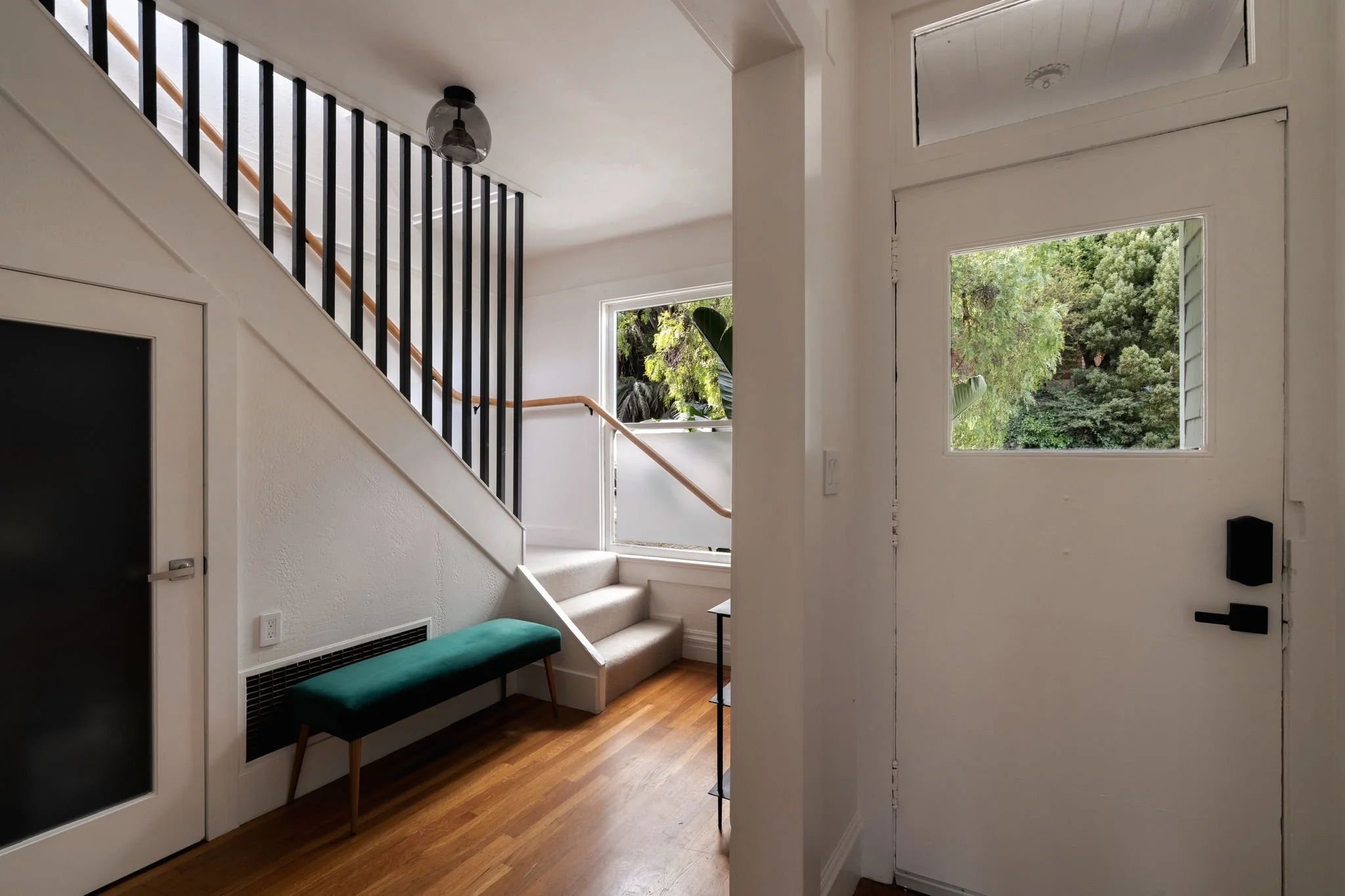 Interior view of a modern home entryway with white walls, hardwood flooring, and a staircase with black railings. Large windows provide natural light, and there is a green cushioned bench near the door.