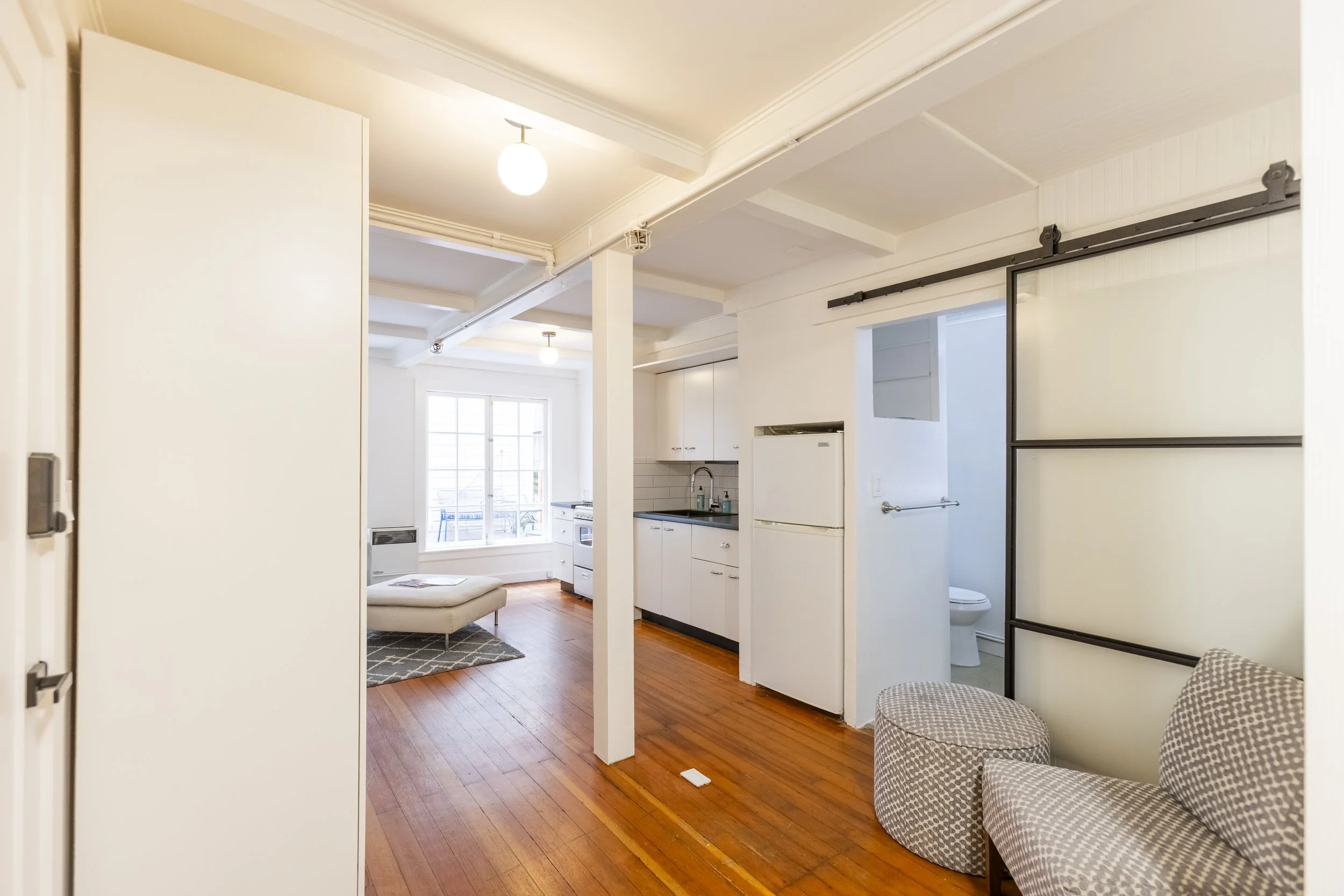 Interior view of a small living space with white walls, hardwood floors, a white couch, and a kitchen area with white cabinets, a small fridge, and a window with natural light.