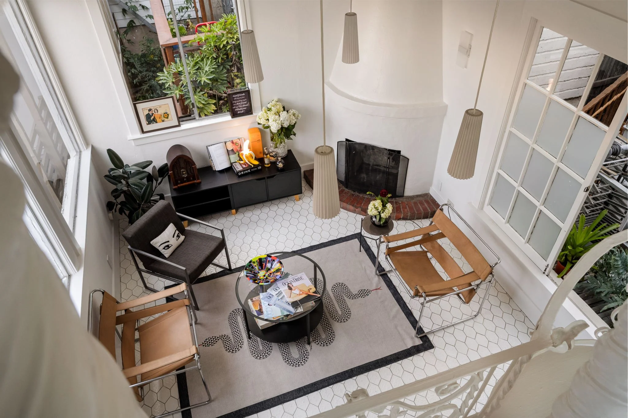 A living room with white walls and hexagonal tile flooring. There are two brown wooden chairs, a gray armchair with a pillow, and two round tables with magazines and colorful decorations. A black media console holds books and decorative items, and fl