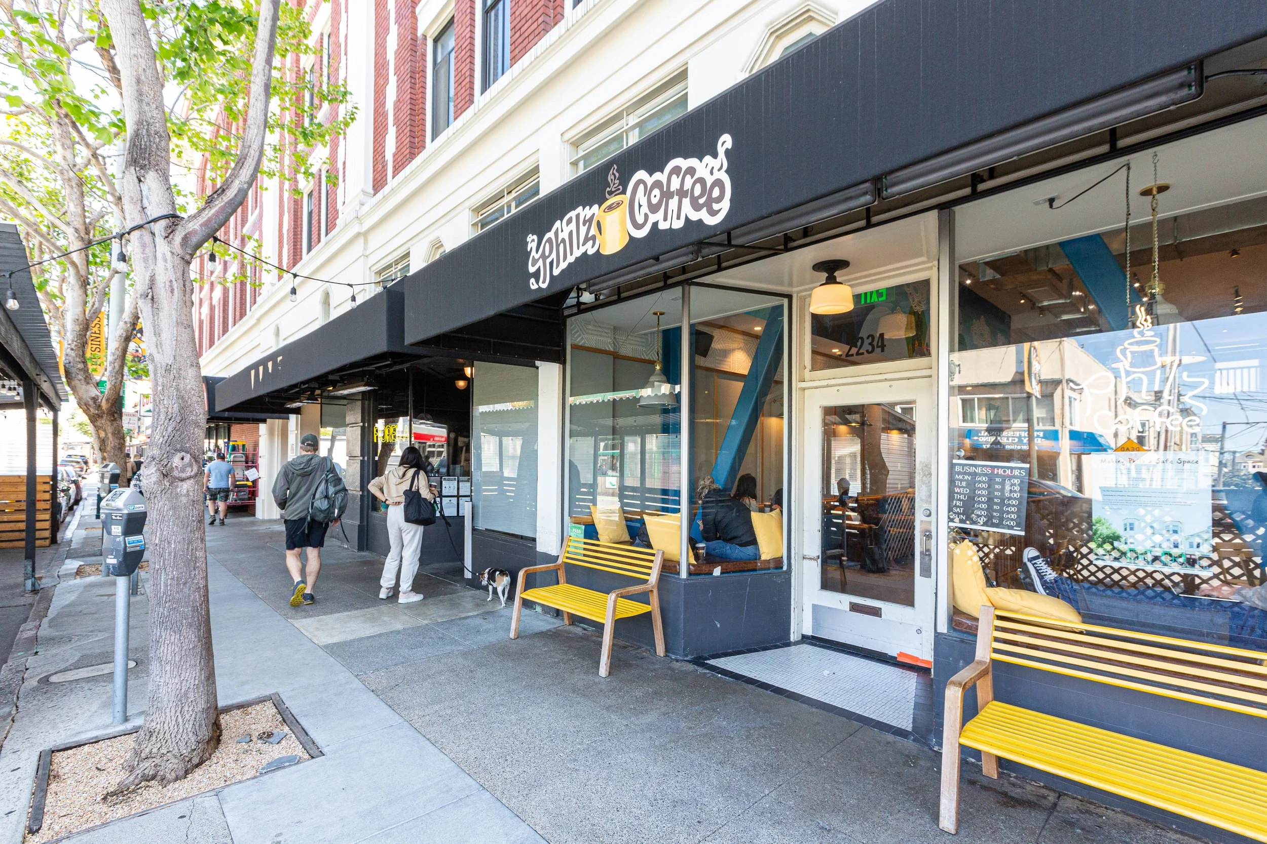 Street view of a coffee shop with yellow benches outside and people walking on the sidewalk.”}
