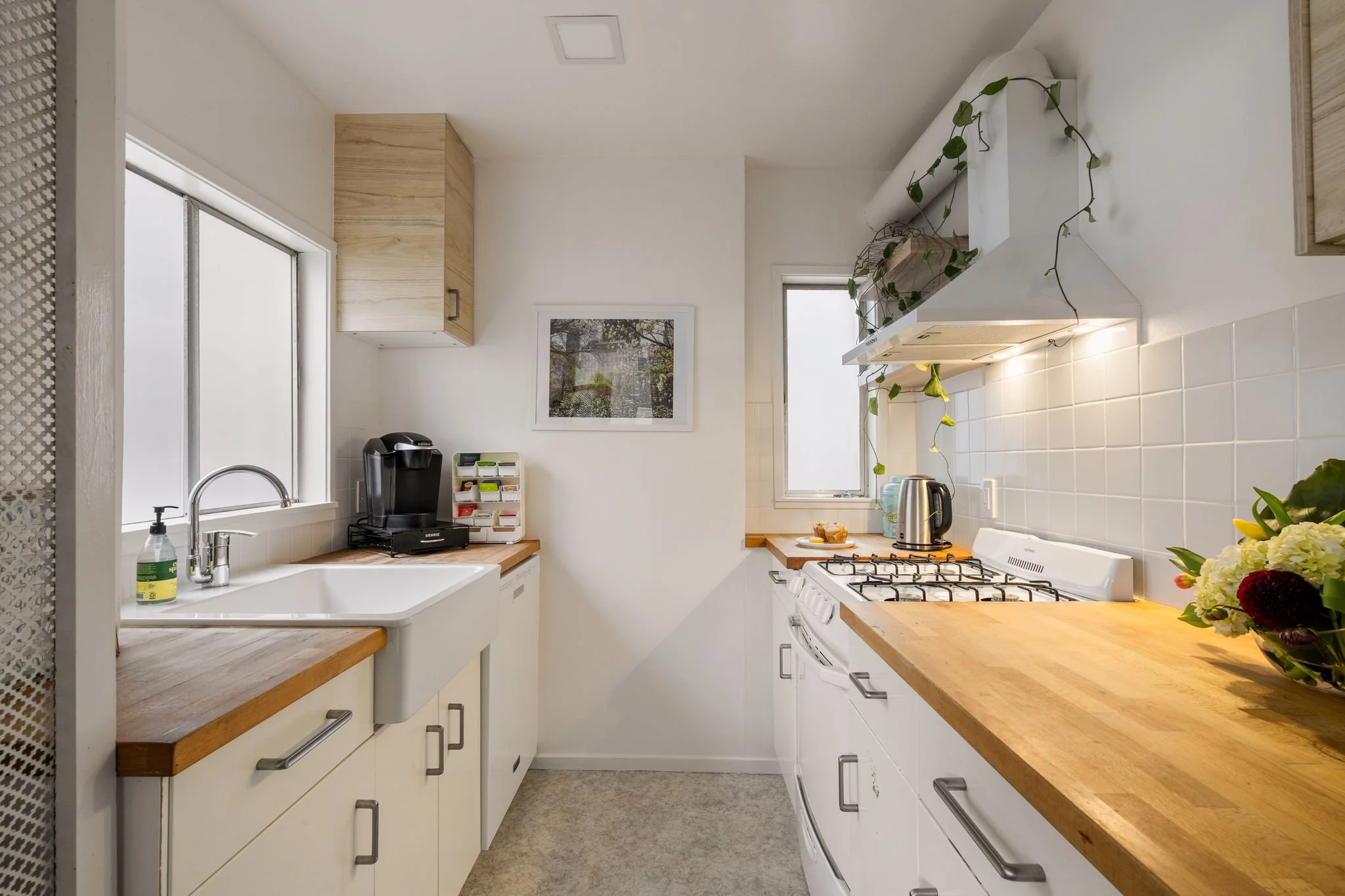 A cozy kitchen with white cabinets, wooden countertops, a stainless steel oven, and a gas stove. Two small windows let in natural light. There are potted plants on the range hood and a framed picture on the wall. A coffee maker, a small white storage