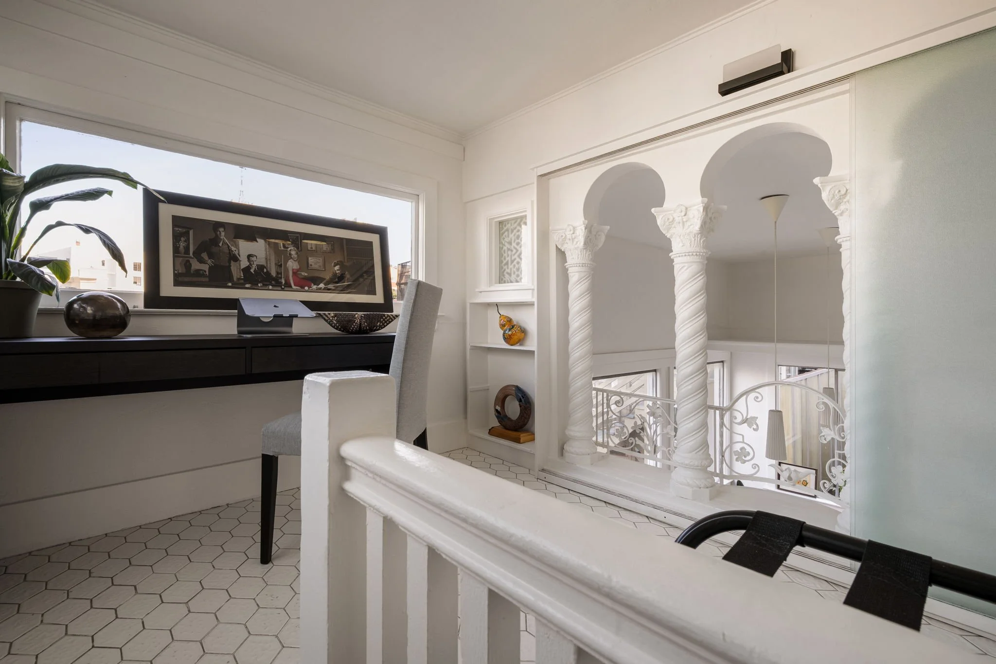 A white balcony railing with a black strap attached to it, overlooking an indoor space with white walls, white columns with decorative capitals, and a hexagonal tile floor. In the background, there is a white cabinet with decorative vases, a dining a