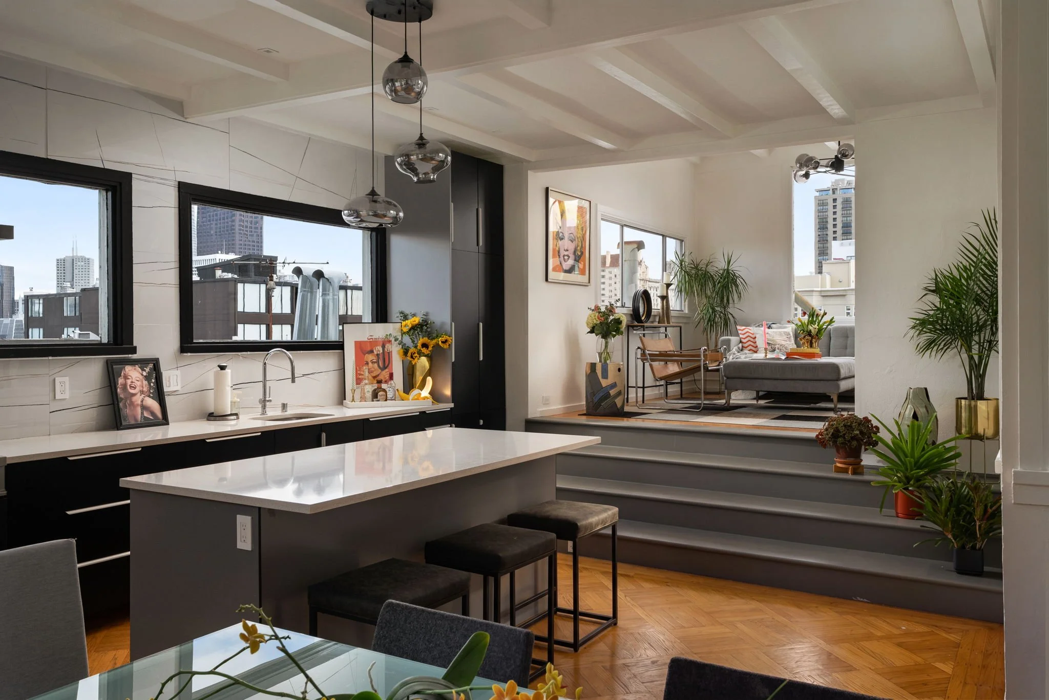 A modern open-concept condo interior with a kitchen and living room, featuring large windows with city skyline views, contemporary artwork, and various decorative plants.