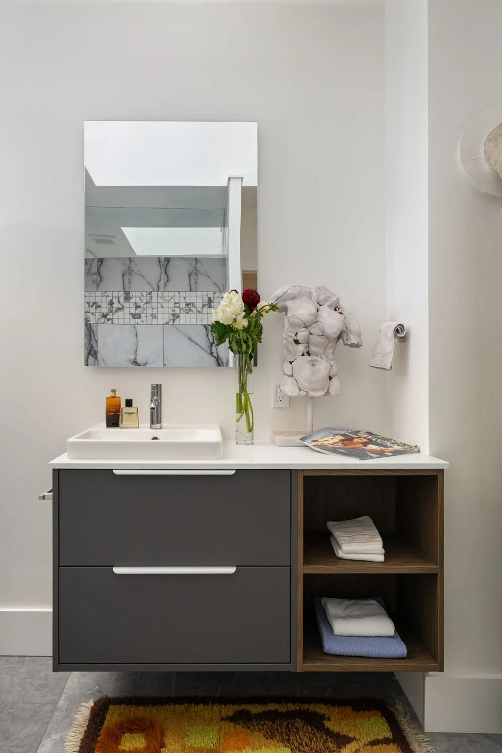 Modern bathroom vanity with a white sink, gray cabinet, wooden open shelves with folded towels, a large mirror, a vase with flowers, and decorative items.