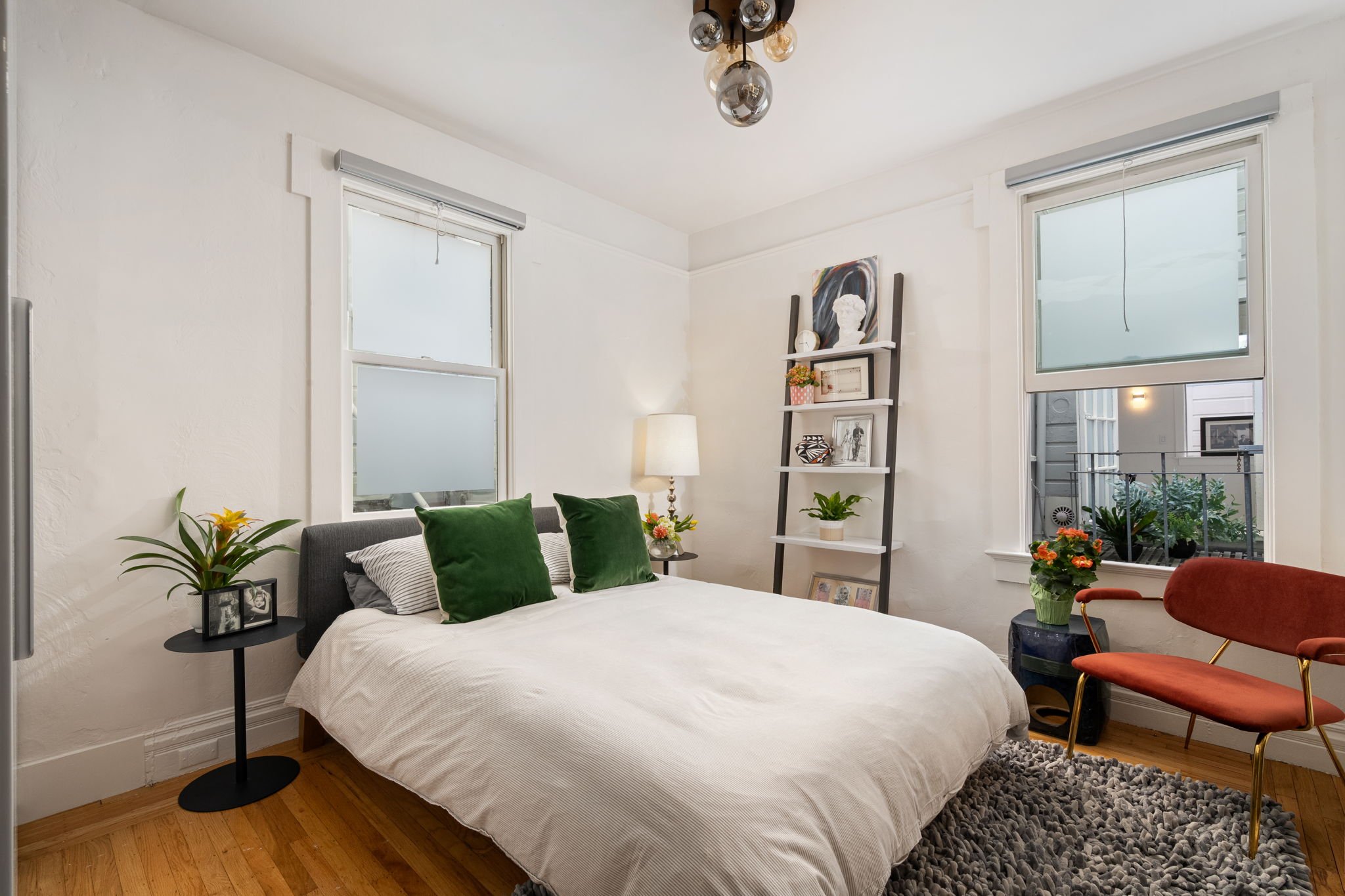 A bright bedroom with white walls and two large windows. The bed has a white cover with two green pillows. Next to the bed are small black side tables with potted plants and framed photos. A tall black ladder-style shelf holds decorative items, inclu