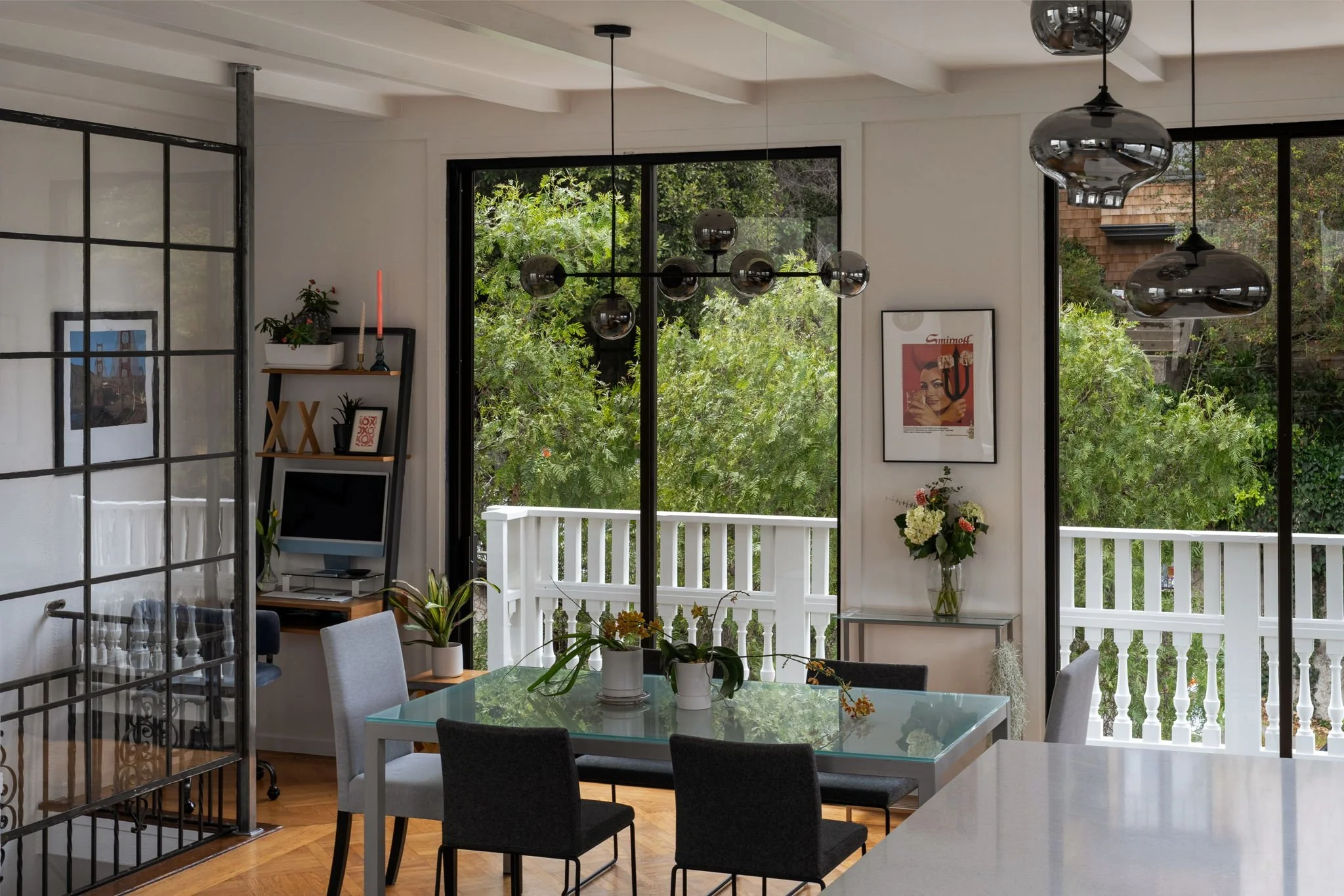 Modern interior dining area with glass-top table, six chairs, potted plants, large windows showing greenery outside, black framed decor, and pendant light fixtures.